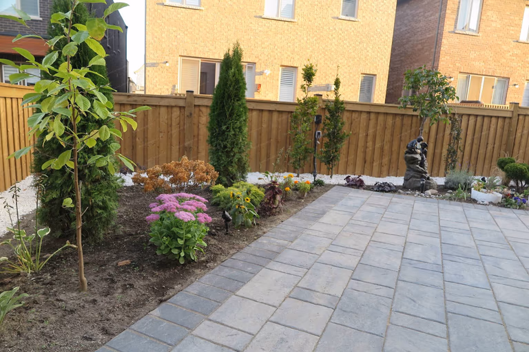 This Vaughan landscaping renovation features a modern interlock patio combined with a carefully arranged garden bed, decorative river rocks, and ornamental plants. The design integrates natural textures, structured greenery, and landscape accents to create a relaxing and visually balanced backyard environment. Professionally completed by Reno Compass Inc.