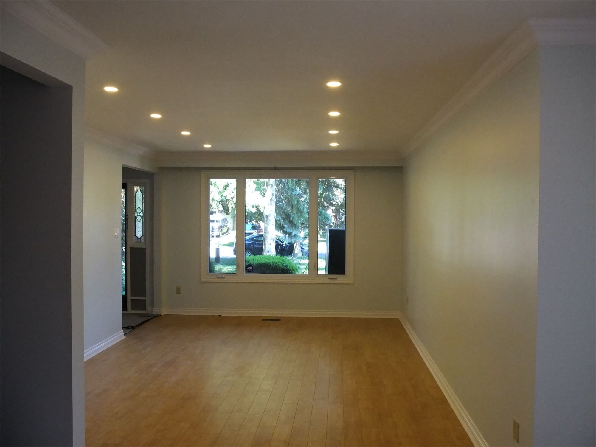 This Oshawa interior renovation highlights a fully refreshed living room with upgraded recessed pot lights, smooth ceiling finish, and newly refinished hardwood flooring. The large front-facing window enhances natural daylight, creating a bright and welcoming atmosphere. Neutral wall tones combined with detailed crown molding provide a clean and modern aesthetic. The optimized lighting layout improves both functionality and visual depth of the space. Completed by a professional Toronto Renovation Company, this project demonstrates quality craftsmanship and thoughtful interior enhancement.