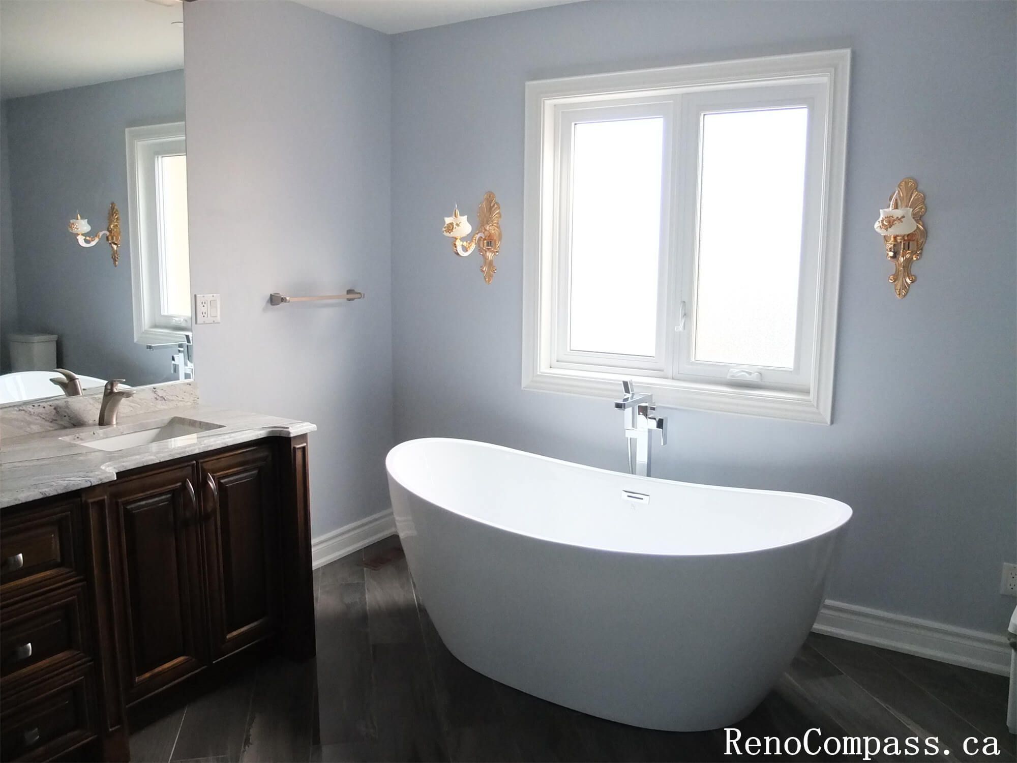 This Richmond Hill interior renovation features a bright and elegant bathroom upgrade with a freestanding soaking bathtub, custom wood vanity, and quartz countertop. Large windows bring in natural light, enhancing the calm and luxurious atmosphere of the space. Modern wall lighting, updated fixtures, and durable flooring create a functional yet spa-like environment for everyday comfort. Completed by a trusted Toronto renovation company, this bathroom renovation blends timeless design with high-quality materials to improve both lifestyle and home value.