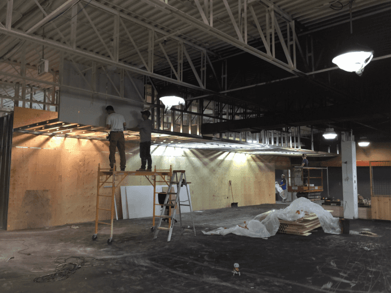 This image captures the construction phase of an outdoor retail store renovation, featuring structural ceiling framing, custom wood wall installation, and lighting preparation. The team is installing overhead framing and wood panel backdrops to support future retail displays and shelving systems. Reno Compass Inc. ensures precise craftsmanship and proper structural alignment during every stage of the commercial renovation process, building a durable and well-designed retail environment from the ground up.