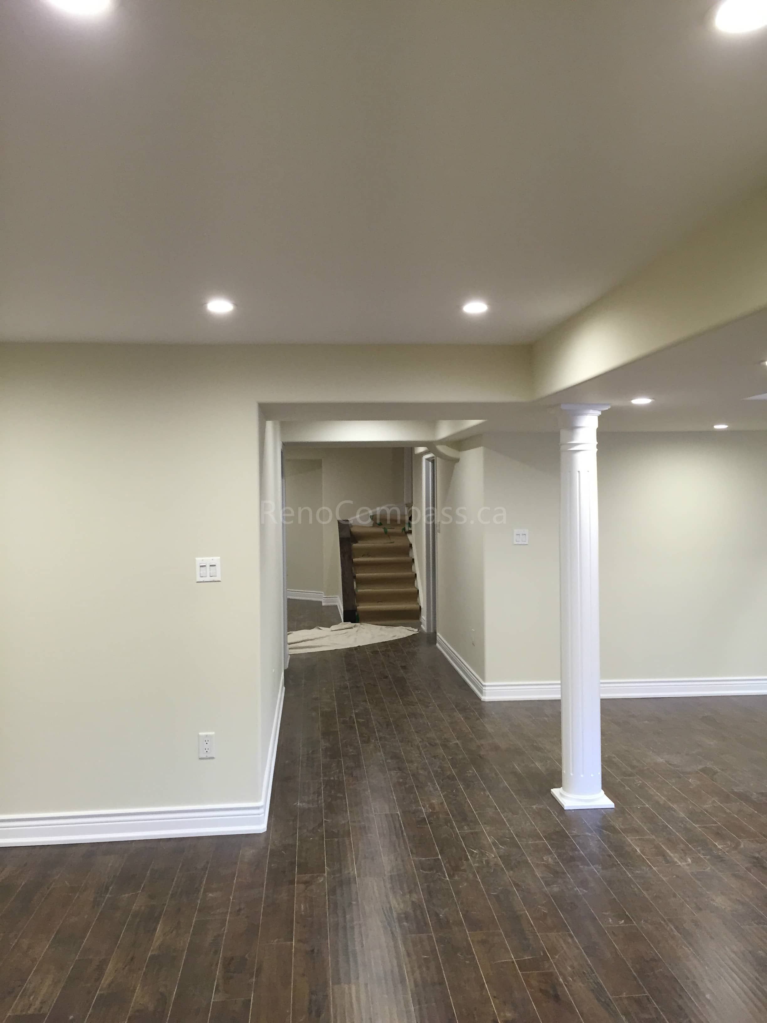 This Toronto interior renovation showcases a newly finished basement with an open-concept layout, dark hardwood flooring, and modern recessed pot lighting. The clean neutral wall tones and structural support columns create a bright and functional living space ideal for entertainment or family use. The renovation enhances both comfort and property value through professional craftsmanship and thoughtful design. Completed by Reno Compass Inc., this project highlights high-quality basement finishing and interior renovation services in Toronto.