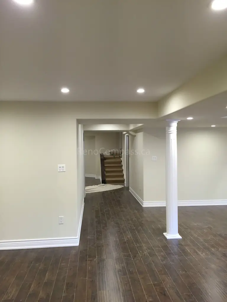 This Toronto interior renovation showcases a newly finished basement with an open-concept layout, dark hardwood flooring, and modern recessed pot lighting. The clean neutral wall tones and structural support columns create a bright and functional living space ideal for entertainment or family use. The renovation enhances both comfort and property value through professional craftsmanship and thoughtful design. Completed by Reno Compass Inc., this project highlights high-quality basement finishing and interior renovation services in Toronto.