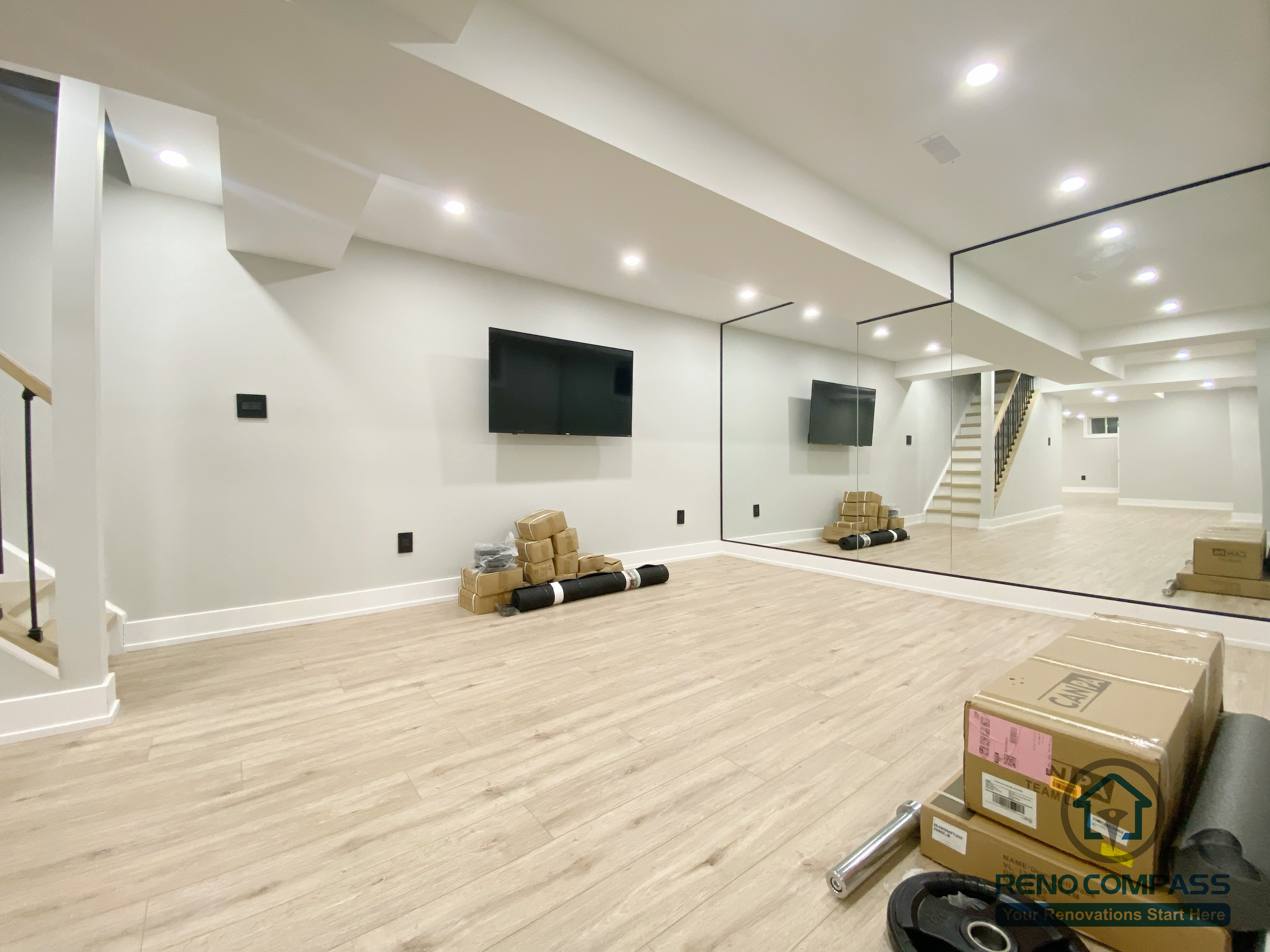 This Aurora basement renovation showcases a spacious entertainment and multi-purpose area featuring a full mirror wall, wall-mounted TV, recessed ceiling lighting, and wood-look flooring. The mirrored surface enhances visual depth and brightness, making the space suitable for home entertainment, fitness, or flexible recreational use. Completed by a Toronto renovation company, this basement renovation highlights adaptable space planning and modern interior finishes.