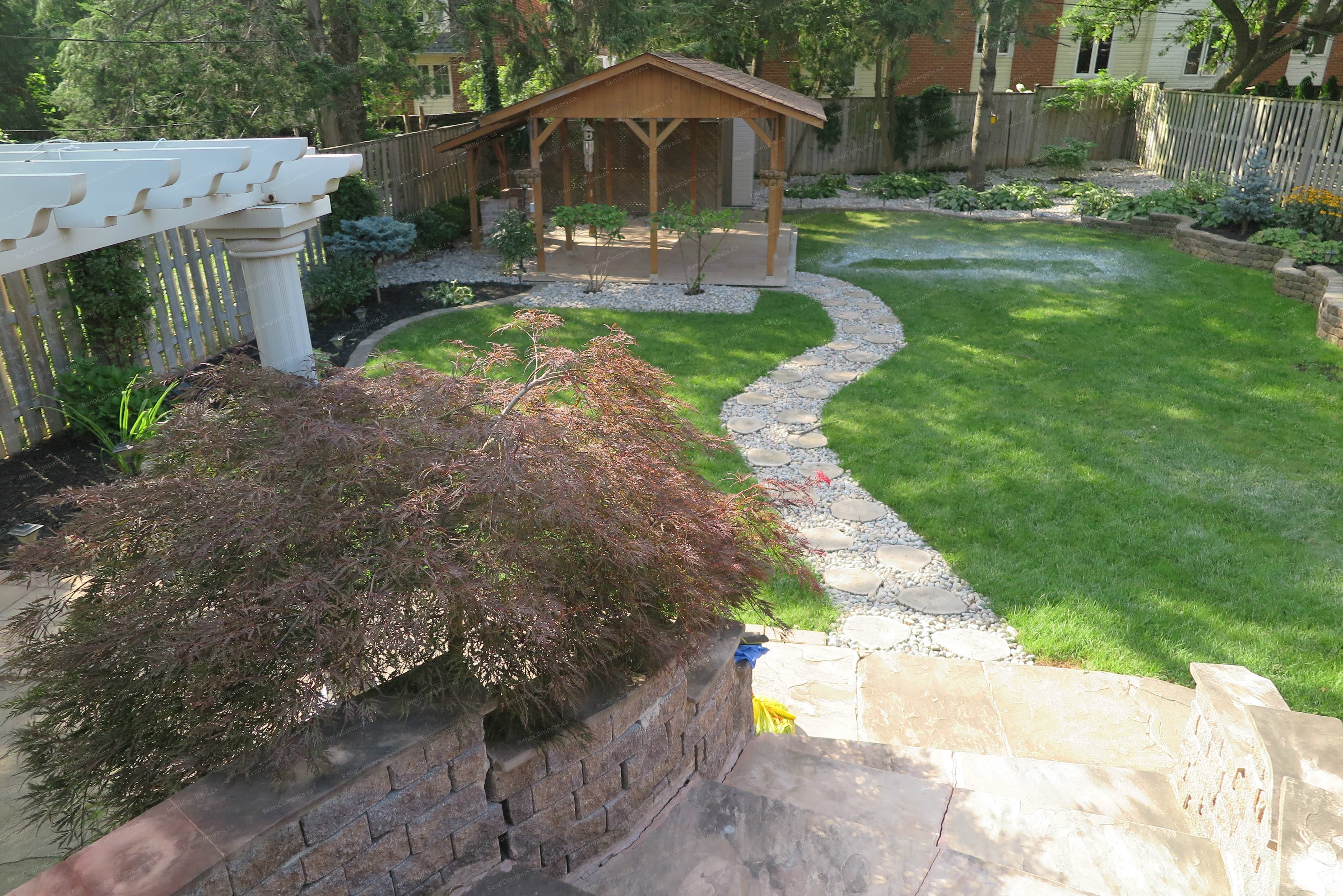 Beautiful North York outdoor landscaping project featuring a custom wooden garden pavilion, curved natural stone walkway, interlock retaining wall, and lush green lawn. Designed and built by Reno Compass to create a relaxing and functional backyard retreat with elegant garden structure and professional landscaping layout.