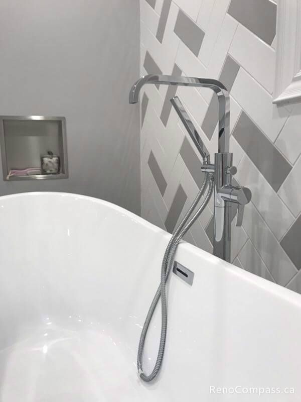 A close-up view of a luxury bathroom renovation in Markham. Showcasing a sleek chrome standing faucet, a white soaking tub, and custom grey and white herringbone wall tiling.