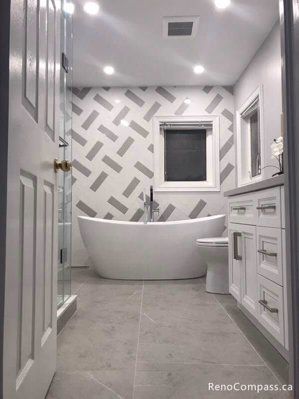 This Markham bathroom renovation features a modern freestanding bathtub, custom white vanity cabinets, quartz countertop, and elegant patterned wall tiles. The renovation includes new flooring tiles, upgraded lighting, and a frameless glass shower, creating a clean and contemporary bathroom design that enhances both comfort and functionality for the homeowner.