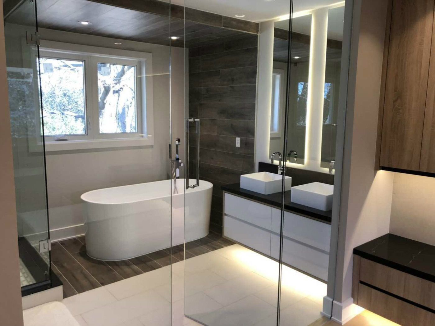 Upscale master bathroom renovation in Markham featuring a freestanding soaking tub, frameless glass shower, double vessel sinks, and custom floating vanity with under-cabinet LED lighting. The combination of wood accent walls, large-format tiles, and modern lighting creates a spa-inspired retreat that blends warmth, elegance, and everyday functionality for contemporary homes.