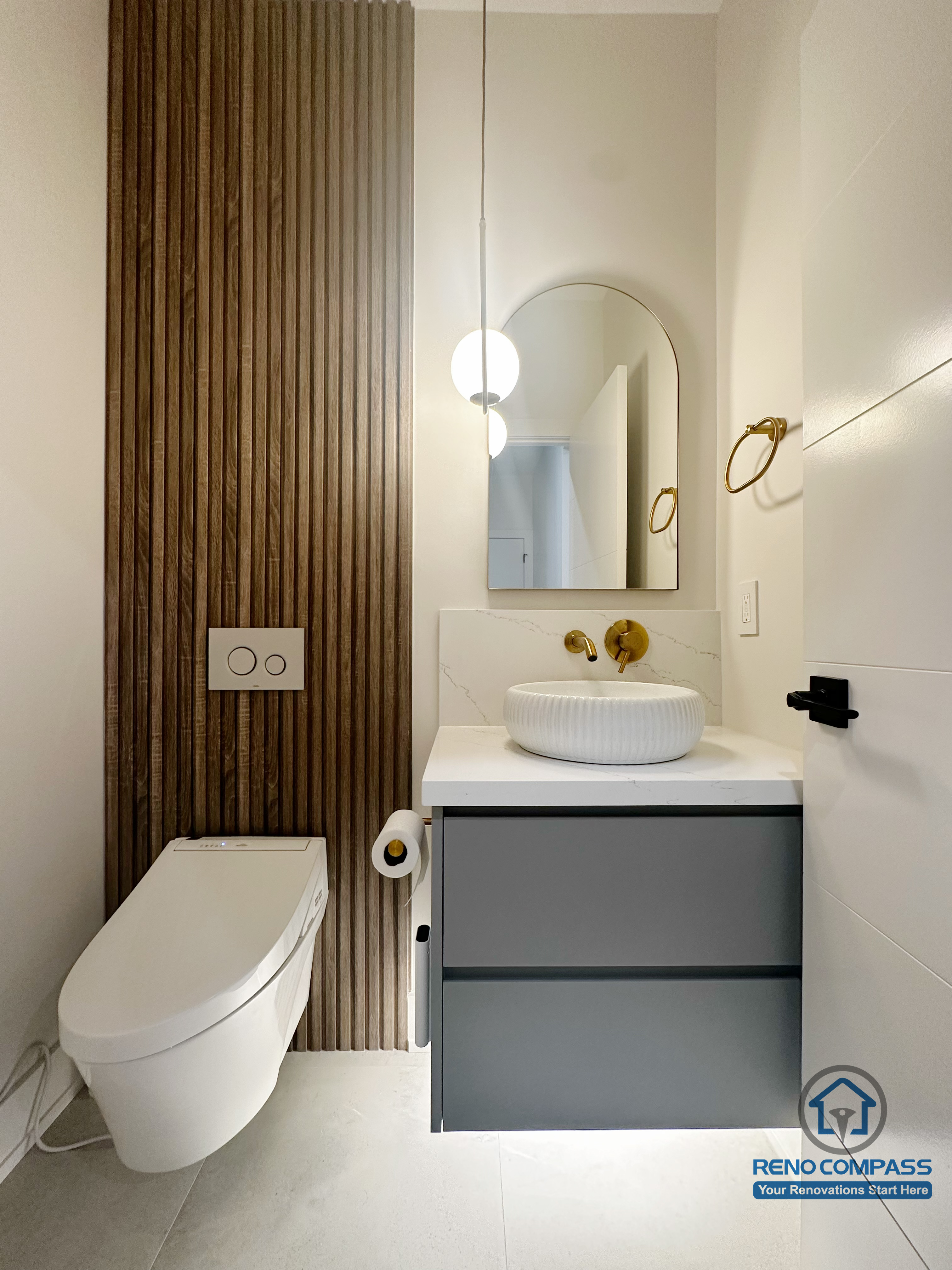 This Oakville powder room renovation features a modern floating vanity paired with a vessel sink and warm brass fixtures. A vertical wood slat accent wall adds texture and contrast, while the wall-mounted toilet and compact layout maximize visual openness, creating a refined and functional small-space bathroom design.