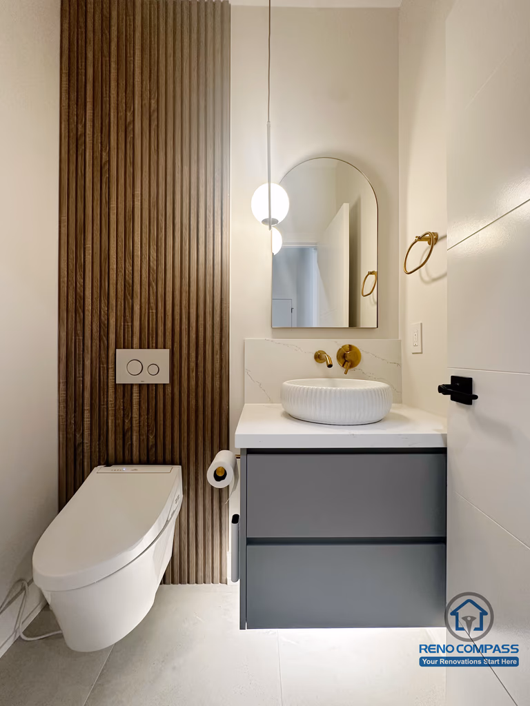 This Oakville powder room renovation features a modern floating vanity paired with a vessel sink and warm brass fixtures. A vertical wood slat accent wall adds texture and contrast, while the wall-mounted toilet and compact layout maximize visual openness, creating a refined and functional small-space bathroom design.