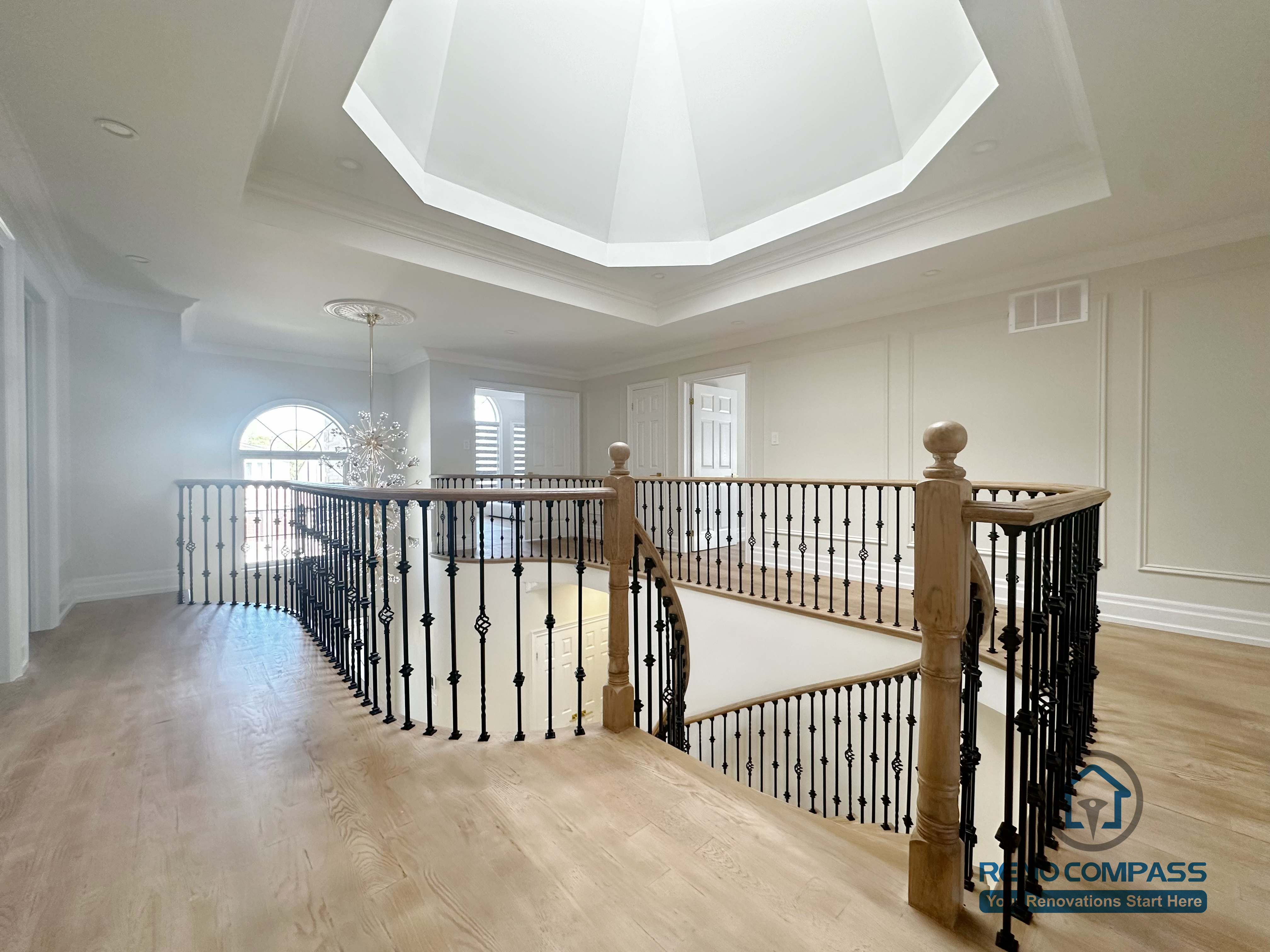 This image showcases a combined flooring and stair renovation completed in North York, featuring hardwood flooring on the upper level and a curved stair railing overlooking the main floor. The staircase is framed with decorative iron balusters and a smooth hardwood handrail, creating continuity between levels. Natural light from the overhead skylight enhances the warmth of the flooring and highlights the architectural detailing. This renovation demonstrates seamless coordination between flooring installation and stair design, improving both visual flow and spatial connection within the home.