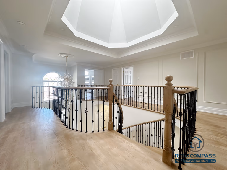 This image showcases a combined flooring and stair renovation completed in North York, featuring hardwood flooring on the upper level and a curved stair railing overlooking the main floor. The staircase is framed with decorative iron balusters and a smooth hardwood handrail, creating continuity between levels. Natural light from the overhead skylight enhances the warmth of the flooring and highlights the architectural detailing. This renovation demonstrates seamless coordination between flooring installation and stair design, improving both visual flow and spatial connection within the home.