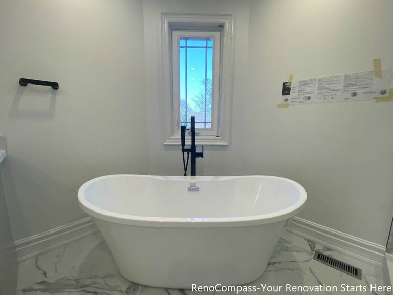 This Thornhill bathroom renovation features a freestanding bathtub positioned beneath a window, creating a calm and balanced focal point within the space. The tub is paired with a matte black floor-mounted faucet, adding a modern contrast to the light marble-look floor tiles. Natural daylight enhances the clean, minimal design, while the open layout promotes comfort, relaxation, and long-term functionality for everyday use.