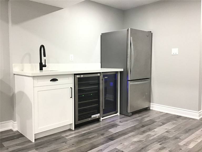 Elegant basement wet bar installation in this Newmarket renovation featuring white shaker cabinetry, quartz countertop, matte black faucet, built-in beverage fridge, and stainless steel refrigerator. A perfect addition for entertaining guests and enhancing basement functionality.