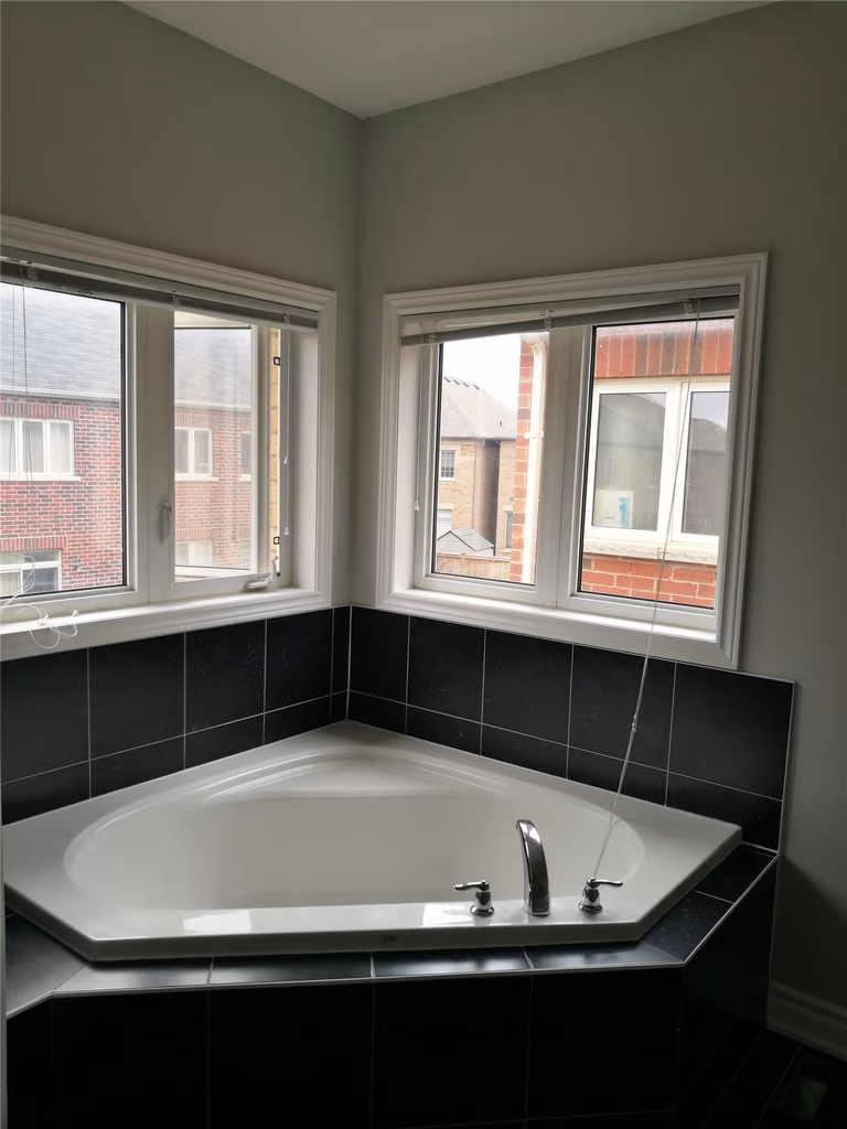 This image features a bathtub area after a Markham interior renovation, showcasing a corner soaking tub, tiled surround, upgraded fixtures, and dual windows that bring in natural light and improve the overall bathroom experience.