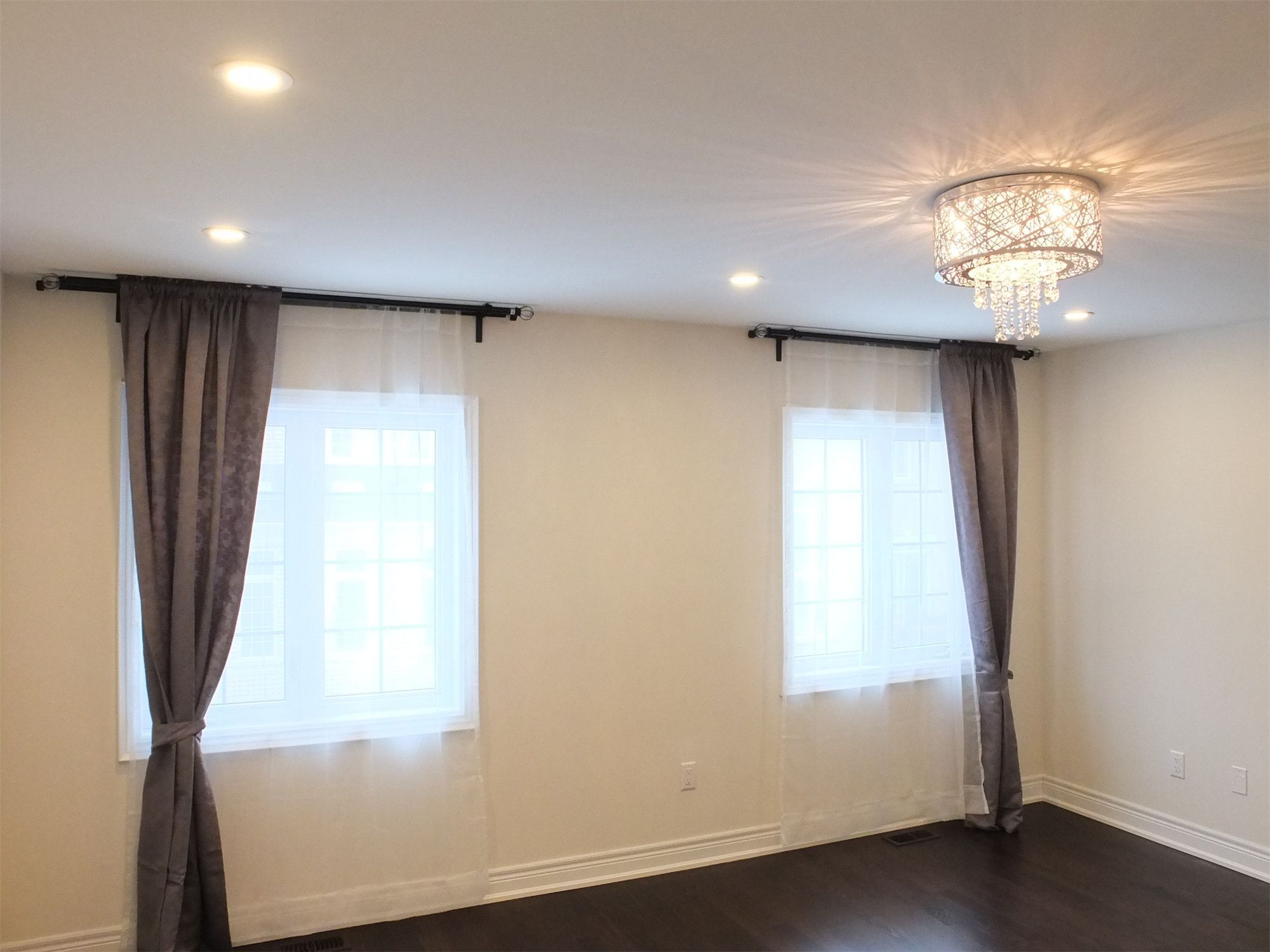 This Kleinburg interior renovation showcases a spacious bedroom featuring elegant crystal ceiling lighting complemented by recessed pot lights for balanced illumination. Large windows with soft curtains allow natural light to brighten the room, while dark hardwood flooring adds contrast and warmth. The combination of modern lighting, neutral wall finishes, and clean architectural lines creates a comfortable and refined living space.