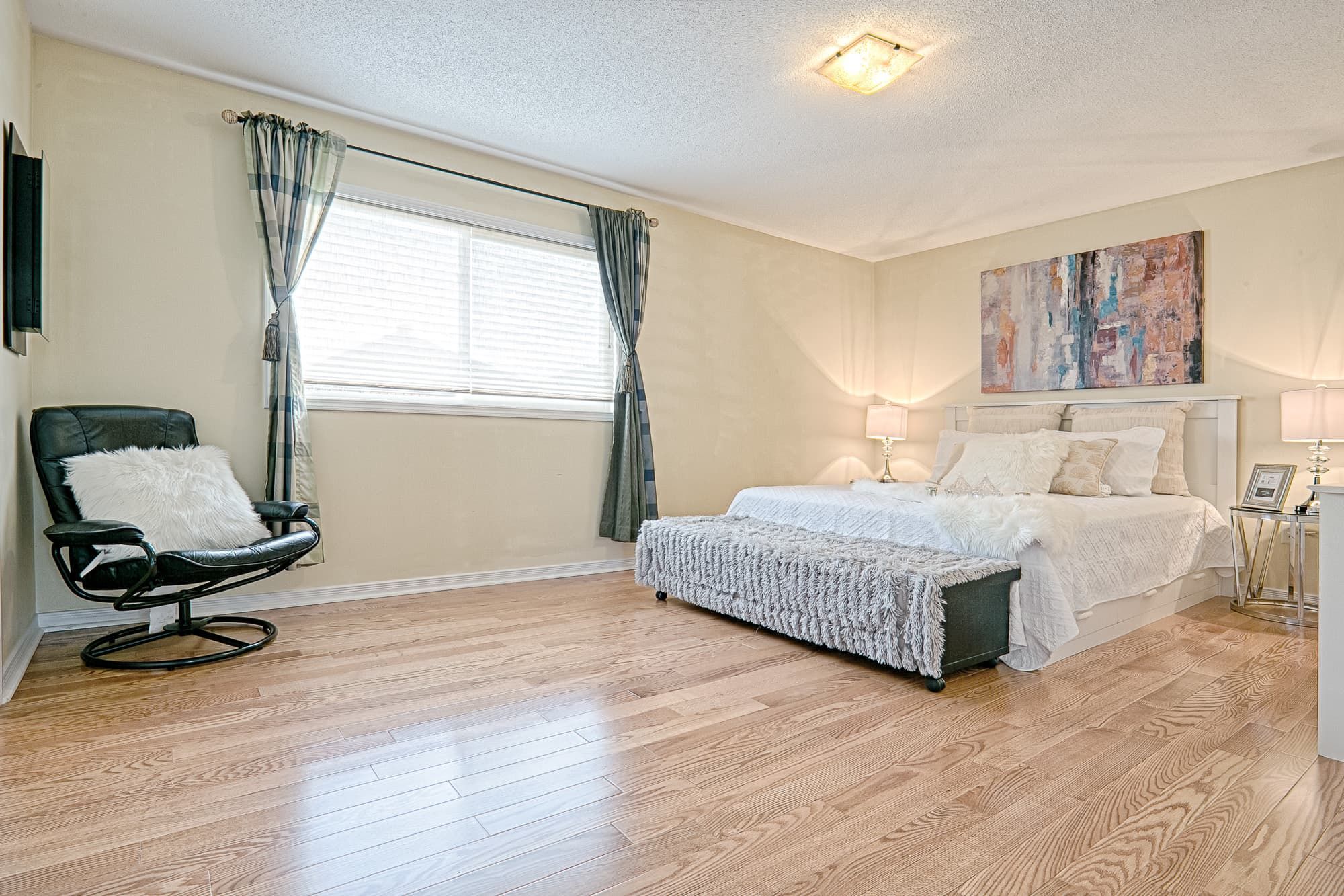 This spacious master bedroom features a simple and lovely interior design with natural hardwood flooring, soft neutral wall tones, and a well-balanced furniture layout. Symmetrical bedside lighting and minimal decor create a clean, relaxing atmosphere, while large windows provide ample natural light. The design focuses on comfort, openness, and timeless style, making it suitable for family homes and long-term residential living.