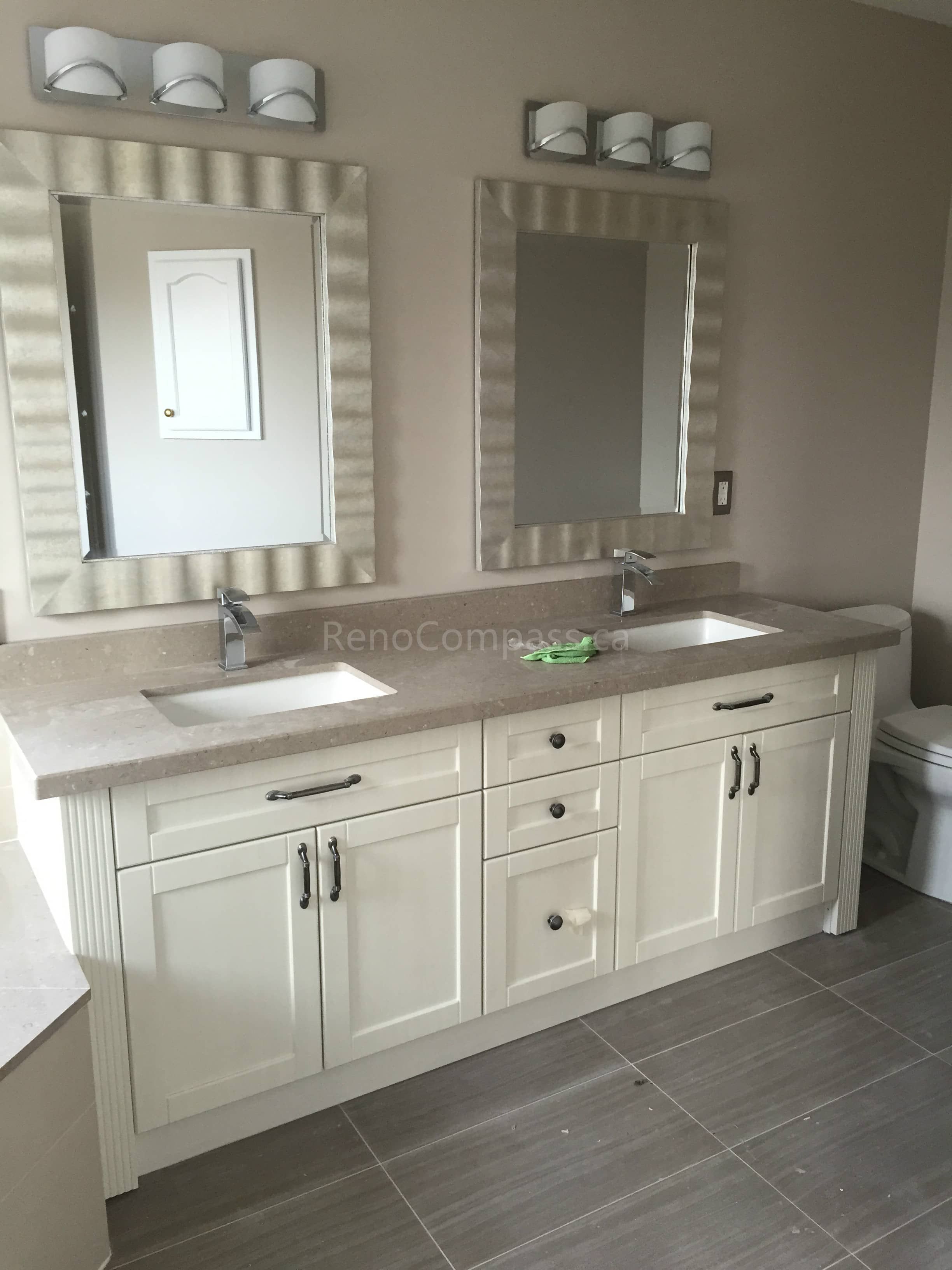 This Toronto bathroom renovation showcases a custom double sink vanity with shaker-style cabinetry and a light quartz countertop. The elegant framed mirrors and modern wall-mounted lighting fixtures enhance the brightness and symmetry of the space. Neutral floor tiles complement the soft cabinetry tones, creating a clean and timeless bathroom design. Completed by Reno Compass Inc., this project highlights quality craftsmanship and functional luxury in Toronto interior renovations.