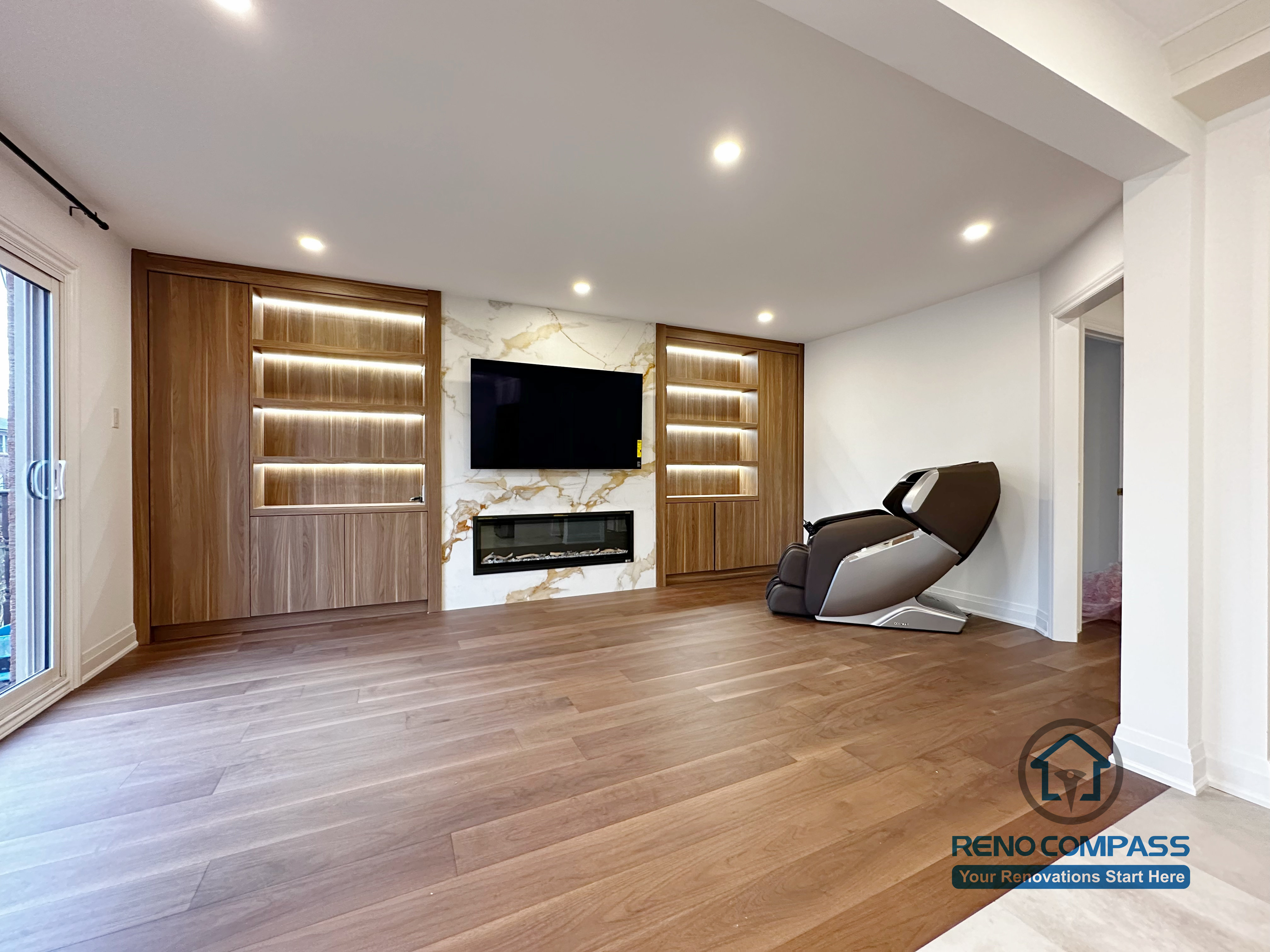 This image highlights a custom-built feature wall completed as part of a Markham interior renovation project. The design features warm wood paneling, integrated LED-lit display shelving, and a marble-look TV backdrop with a modern linear fireplace. Wide-plank hardwood flooring and recessed ceiling lights create a clean, open feel, while the built-in elements add both storage and visual focus. This space was designed to seamlessly connect with the adjacent kitchen and dining areas, reflecting the cohesive craftsmanship of a professional Toronto renovation company.