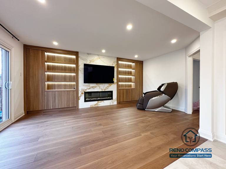 This image highlights a custom-built feature wall completed as part of a Markham interior renovation project. The design features warm wood paneling, integrated LED-lit display shelving, and a marble-look TV backdrop with a modern linear fireplace. Wide-plank hardwood flooring and recessed ceiling lights create a clean, open feel, while the built-in elements add both storage and visual focus. This space was designed to seamlessly connect with the adjacent kitchen and dining areas, reflecting the cohesive craftsmanship of a professional Toronto renovation company.