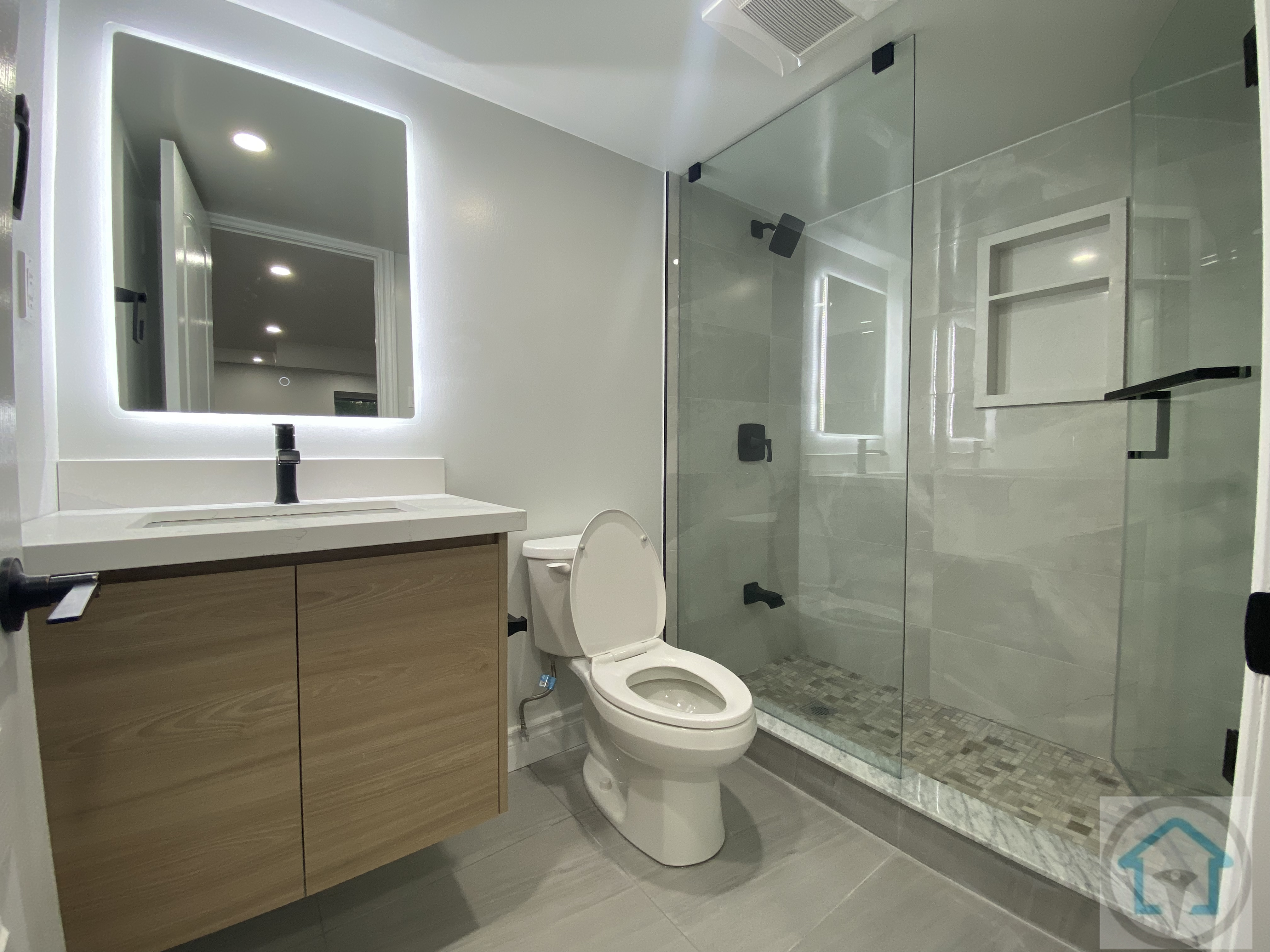 This Aurora basement bathroom renovation features a modern glass shower with marble-style tiles, a sleek wooden vanity, and a contemporary toilet. The LED mirror and recessed lighting enhance the bright and functional space. Contractor: Reno Compass Inc., City: Aurora.