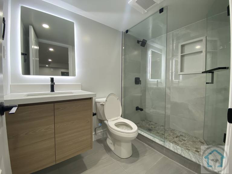 This Aurora basement bathroom renovation features a modern glass shower with marble-style tiles, a sleek wooden vanity, and a contemporary toilet. The LED mirror and recessed lighting enhance the bright and functional space. Contractor: Reno Compass Inc., City: Aurora.