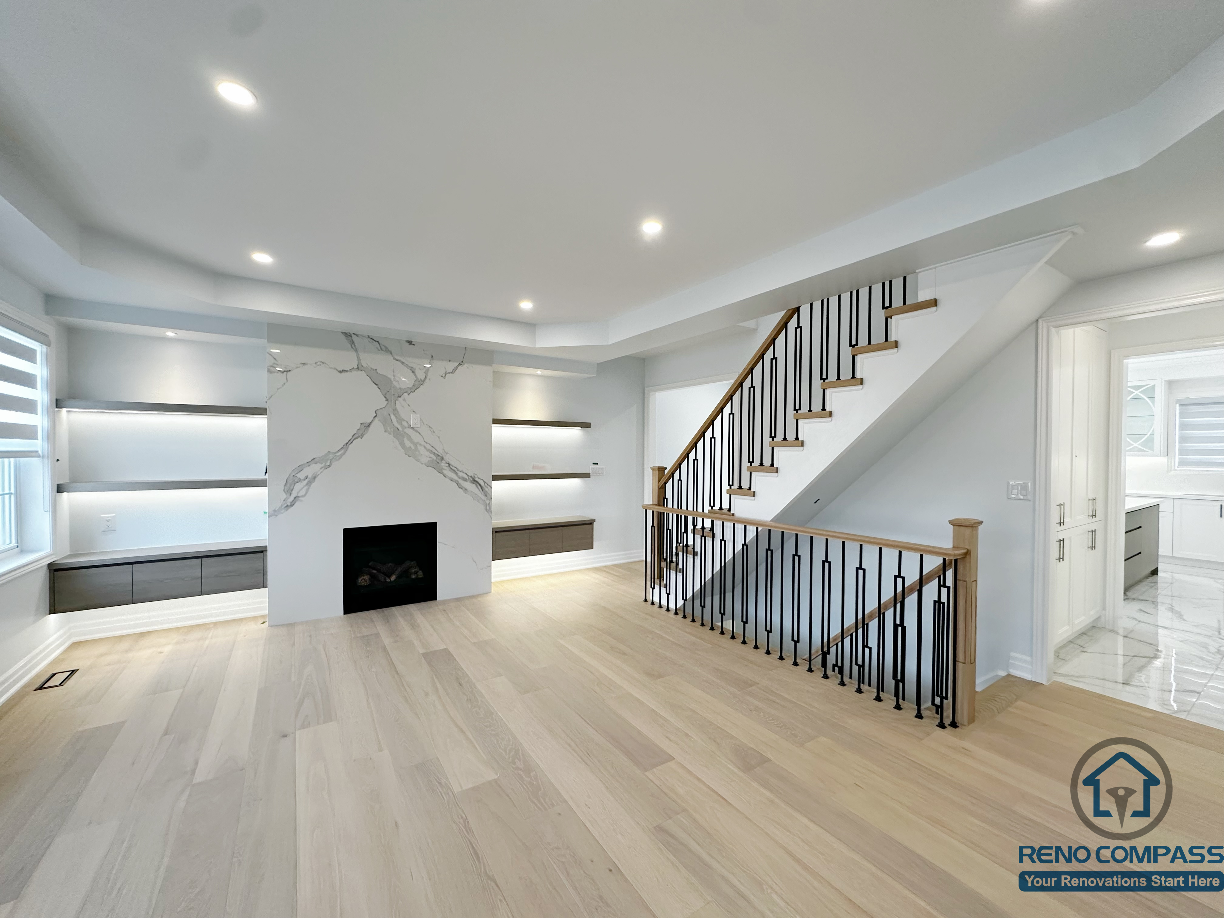 This image showcases an open living area renovation in a Markham house, featuring a custom fireplace wall with marble-style slab detailing, built-in shelving with integrated lighting, and a modern staircase with wood treads and black metal railing. Light-toned hardwood flooring and recessed ceiling lights create a cohesive, bright, and contemporary interior flow.