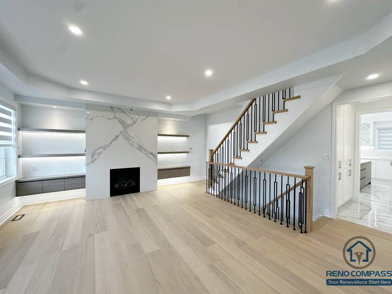 This image showcases an open living area renovation in a Markham house, featuring a custom fireplace wall with marble-style slab detailing, built-in shelving with integrated lighting, and a modern staircase with wood treads and black metal railing. Light-toned hardwood flooring and recessed ceiling lights create a cohesive, bright, and contemporary interior flow.