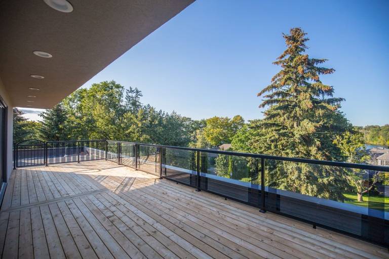 Spacious outdoor terrace of a 5000 sqft Toronto luxury home renovation featuring a wide wooden deck, modern glass railing system, and elevated balcony overlooking mature trees and scenic surroundings. This custom exterior living space enhances outdoor entertaining and relaxation with durable materials and clean contemporary design. Completed by Reno Compass Inc., a trusted Toronto renovation company specializing in high-end residential renovations and full-property transformations.
