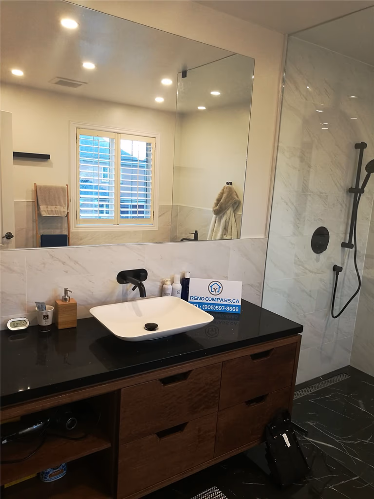 This Richmond Hill bathroom renovation features a contemporary vanity with dark wood cabinetry, a sleek vessel sink, and a large wall-to-wall mirror that enhances light and space. The design is complemented by marble-style wall finishes, black fixtures, and a modern glass shower enclosure, creating a clean and luxurious atmosphere. Completed by a professional Toronto renovation company, this project highlights high-quality materials, practical storage solutions, and timeless modern design for everyday comfort.