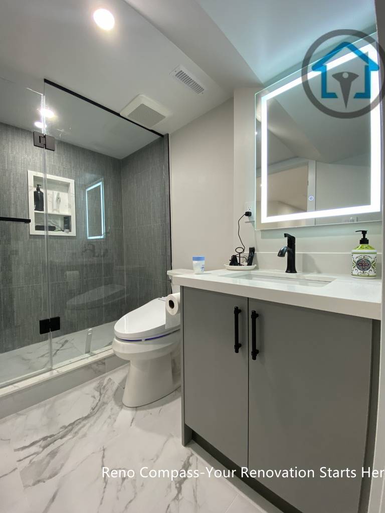 This Stouffville basement bathroom renovation highlights a frameless glass shower, large-format wall tiles, marble-look flooring, LED mirror, and modern vanity with matte black fixtures. The layout maximizes space efficiency while maintaining a clean, contemporary aesthetic, completed by Reno Compass Inc.