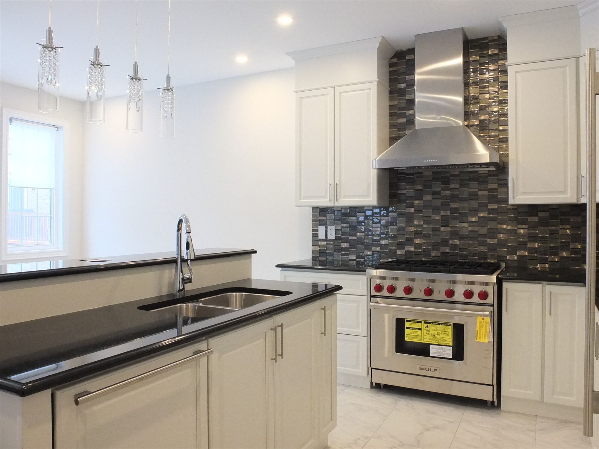 This Kleinburg interior renovation showcases a modern kitchen featuring custom white cabinetry, a spacious island with integrated sink, and sleek black countertops. The kitchen is highlighted by a professional stainless steel gas range and hood, complemented by a dark mosaic tile backsplash that adds depth and contrast. Elegant pendant lighting and large windows enhance the bright, open atmosphere, creating a refined and functional cooking space ideal for modern living.