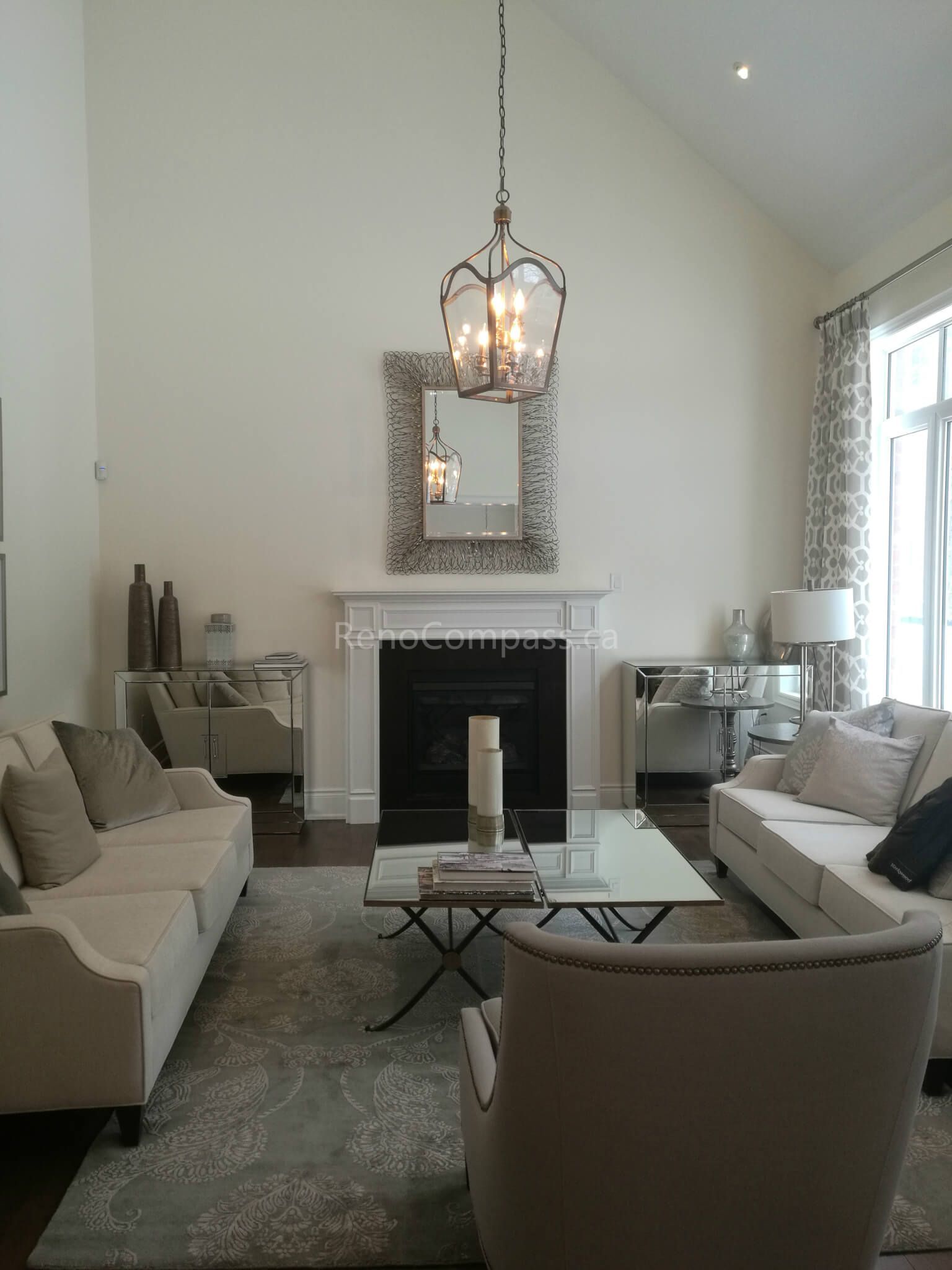 This image features a refined living room completed as part of a Toronto interior renovation project. The space is defined by its high ceiling, central fireplace, and symmetrical furniture layout, creating a balanced and sophisticated atmosphere. Soft neutral tones, layered lighting, and a statement pendant light enhance the sense of height and openness. Large windows allow natural light to brighten the room, while carefully selected furnishings add warmth and comfort. This living room renovation reflects timeless interior design and thoughtful craftsmanship tailored to Toronto residential homes.
