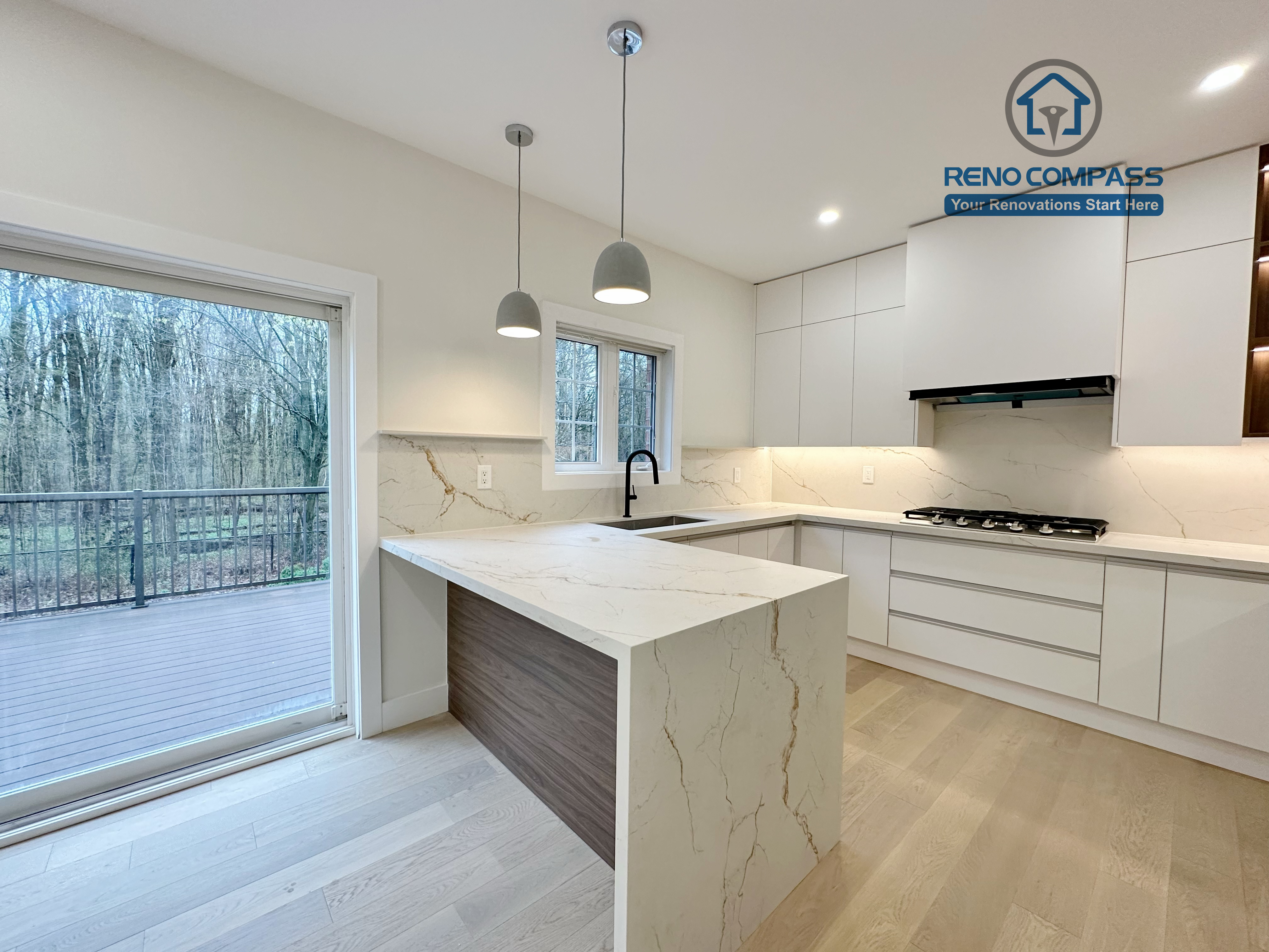 This image presents a modern kitchen renovation in Richmond Hill featuring full-height custom white cabinetry, a quartz waterfall island with wood panel detailing, and integrated stainless steel appliances. The design includes a built-in gas cooktop, minimalist pendant lights, recessed ceiling lighting, and light hardwood flooring for a clean, contemporary look. Large windows and patio access enhance natural light and openness. Completed by a professional Toronto Renovation Company, this kitchen renovation reflects refined craftsmanship, efficient layout planning, and modern family-focused design.