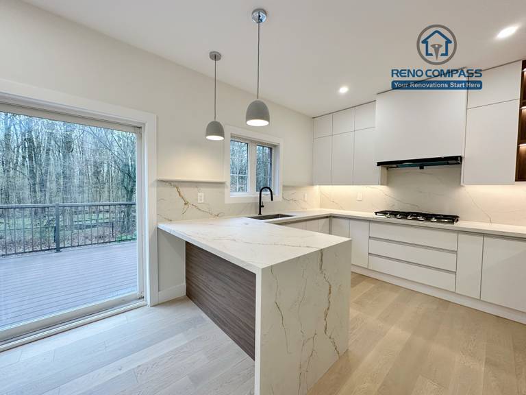 This image presents a modern kitchen renovation in Richmond Hill featuring full-height custom white cabinetry, a quartz waterfall island with wood panel detailing, and integrated stainless steel appliances. The design includes a built-in gas cooktop, minimalist pendant lights, recessed ceiling lighting, and light hardwood flooring for a clean, contemporary look. Large windows and patio access enhance natural light and openness. Completed by a professional Toronto Renovation Company, this kitchen renovation reflects refined craftsmanship, efficient layout planning, and modern family-focused design.