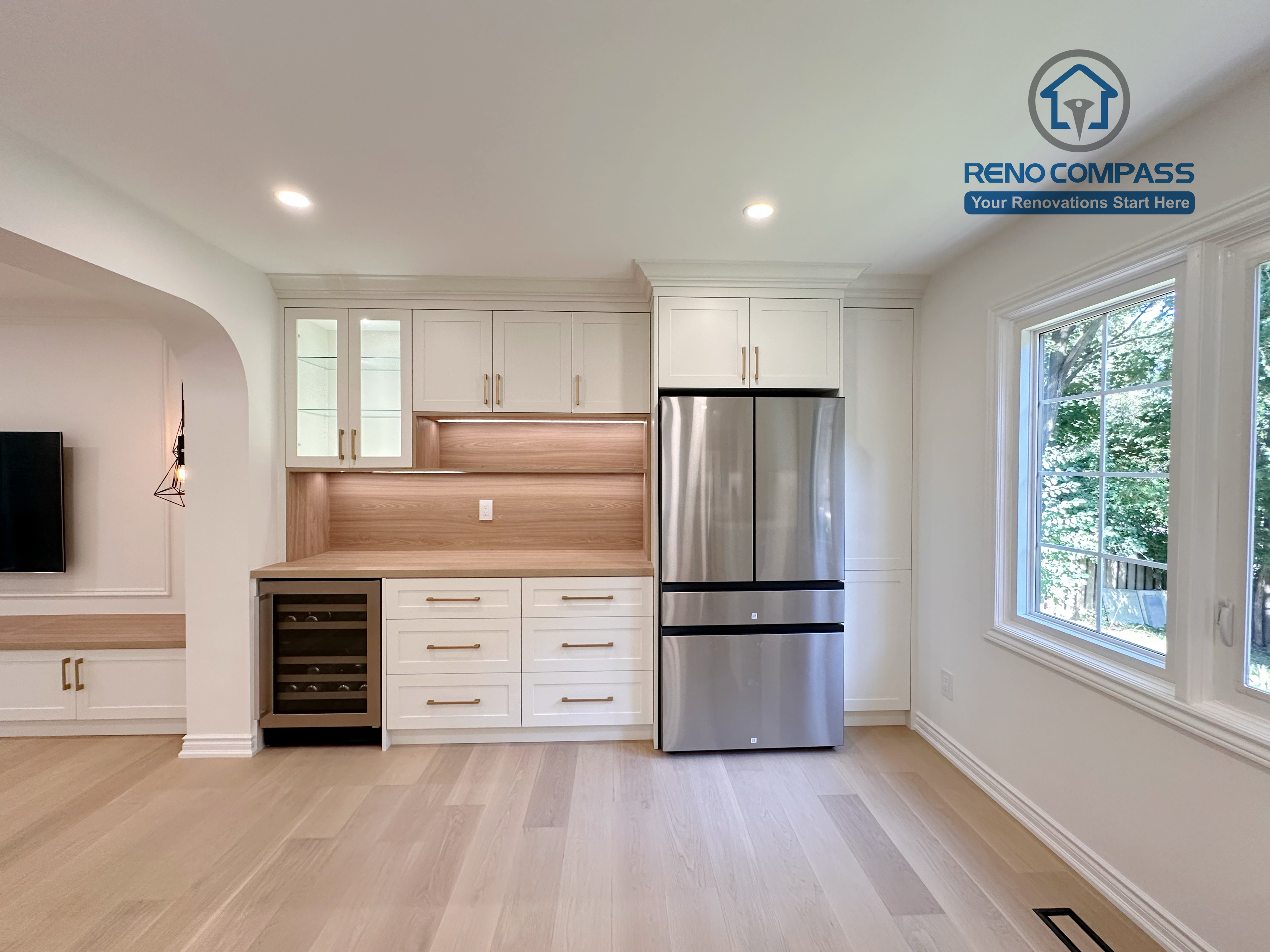 This Markham kitchen renovation features a custom-built appliance wall with full-height cabinetry, integrated storage, and a stainless steel refrigerator. Warm wood backsplash panels and under-cabinet lighting add depth and contrast to the white cabinetry, while large windows bring in natural light. Completed by a professional Toronto renovation company, Reno Compass Inc., the design focuses on organization, balance, and long-term functionality.