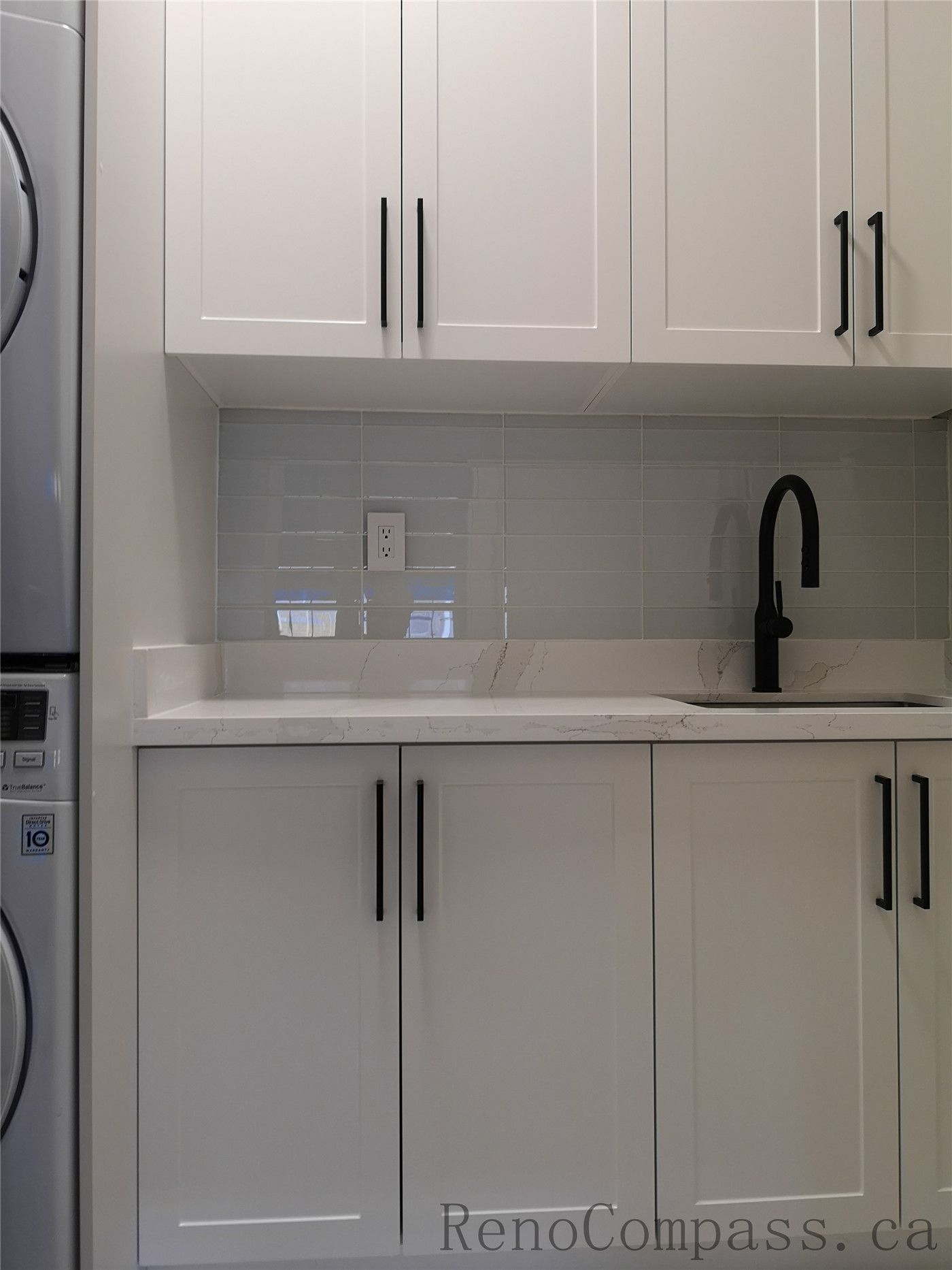 This Aurora home renovation features a clean and modern laundry room design with custom white shaker cabinets, quartz countertop, and a built-in utility sink with matte black faucet. The stacked washer and dryer layout maximizes space efficiency while maintaining a bright and organized look. Completed by a professional Toronto renovation company focused on practical storage solutions, modern finishes, and high-quality craftsmanship.