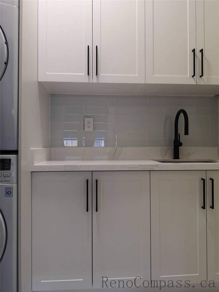This Aurora home renovation features a clean and modern laundry room design with custom white shaker cabinets, quartz countertop, and a built-in utility sink with matte black faucet. The stacked washer and dryer layout maximizes space efficiency while maintaining a bright and organized look. Completed by a professional Toronto renovation company focused on practical storage solutions, modern finishes, and high-quality craftsmanship.