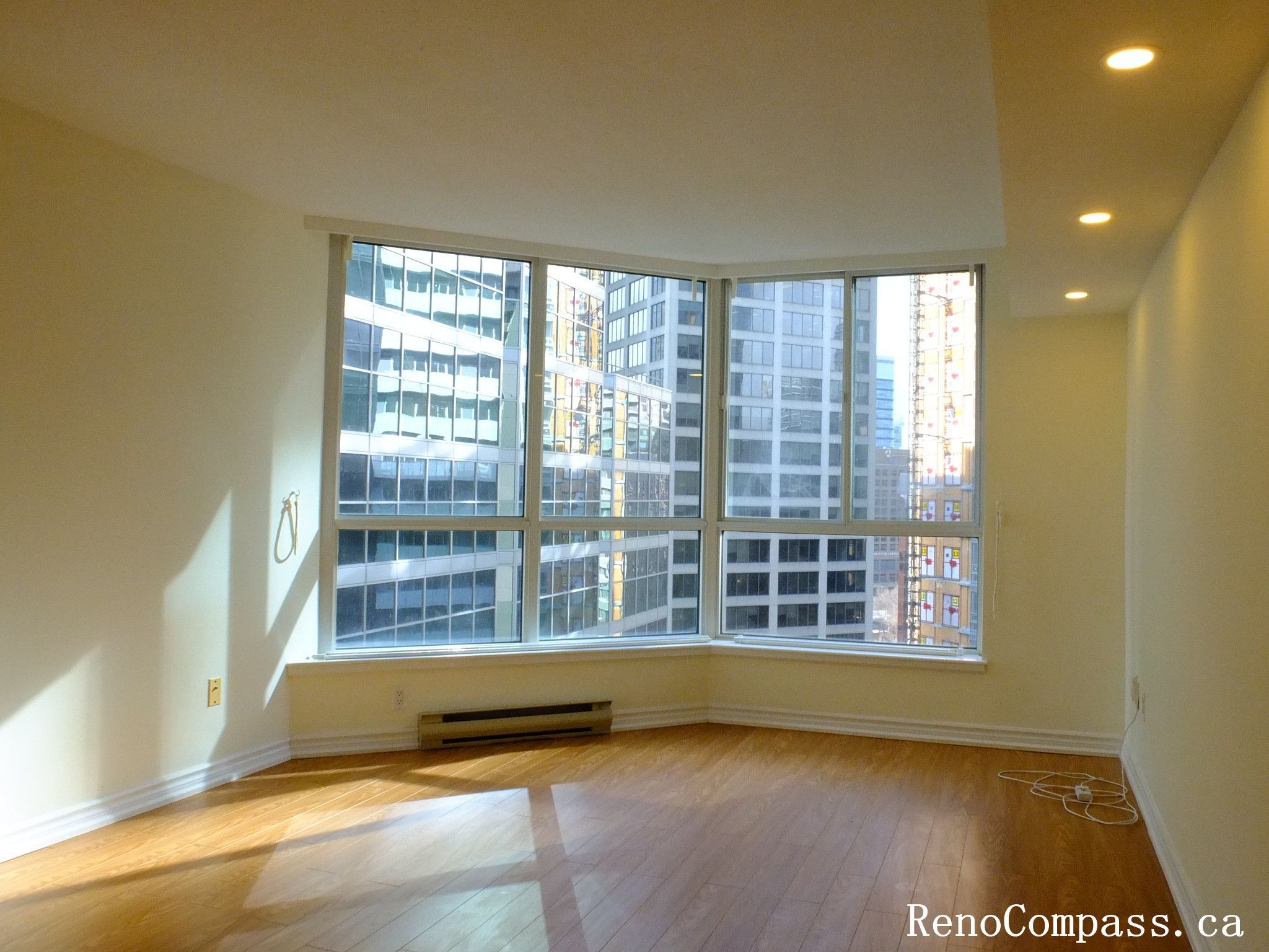 This image shows a bright Toronto condo living room featuring large corner windows with city views and natural sunlight. The space includes hardwood flooring and recessed ceiling lighting, forming part of a Toronto interior renovation project aimed at enhancing brightness, modern design, and overall living comfort.