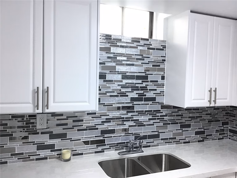 This Whitby basement renovation features a stylish kitchenette area with white shaker cabinets, a double stainless steel sink, and a contemporary glass mosaic backsplash. The neutral quartz-style countertop complements the modern tile design, creating a clean and functional space perfect for entertaining or multi-generational living. Professional cabinet installation and precise tile work enhance both durability and visual appeal.