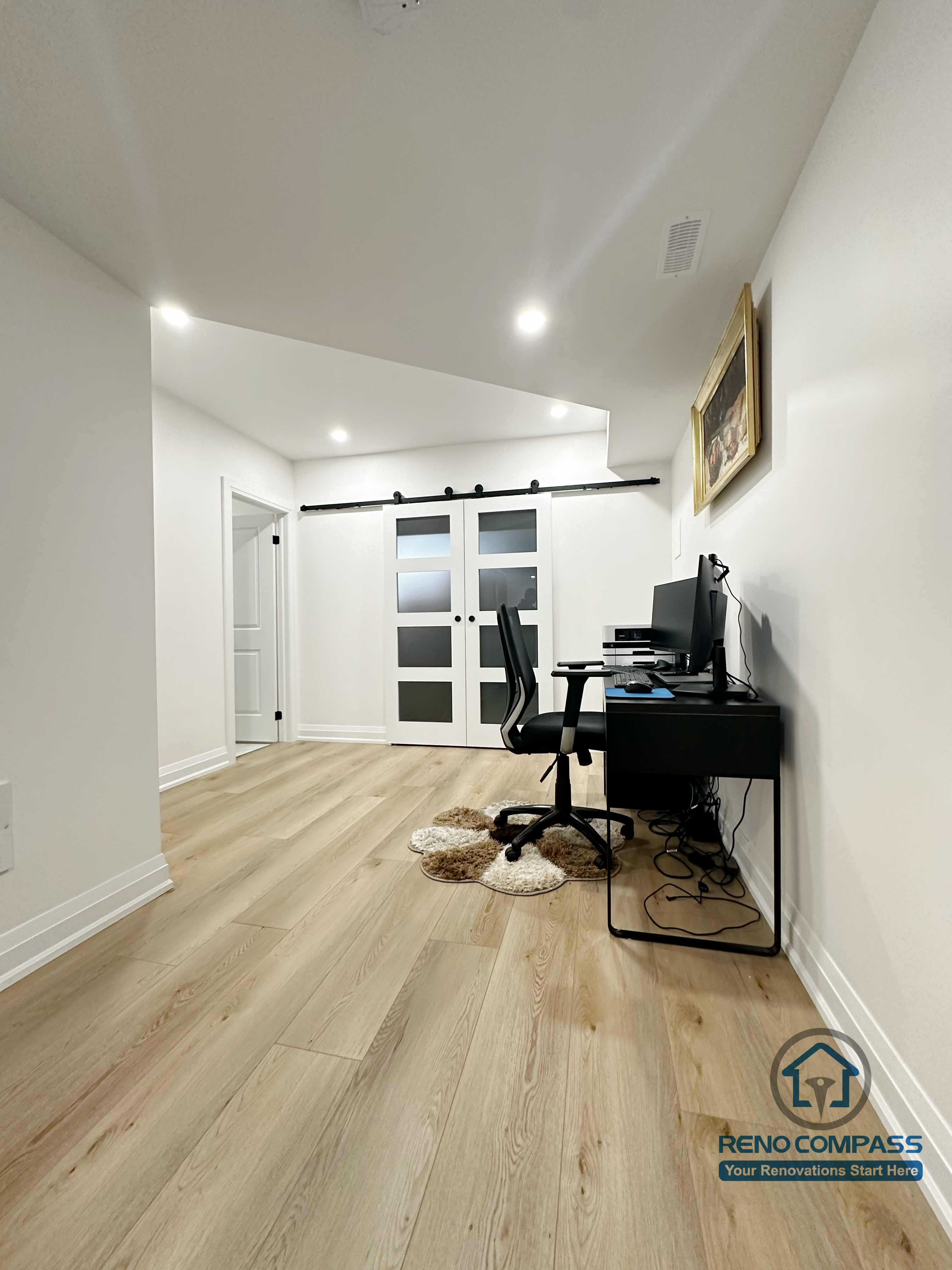 This Richmond Hill basement renovation features a flexible home office area designed for remote work or study. The space includes wide-plank flooring, recessed ceiling lights, and clean white walls that keep the area bright and uncluttered. Sliding barn-style doors add visual interest while allowing separation between work and living zones. The layout comfortably fits a desk setup and can easily adapt into a study room, hobby area, or multi-purpose basement workspace.