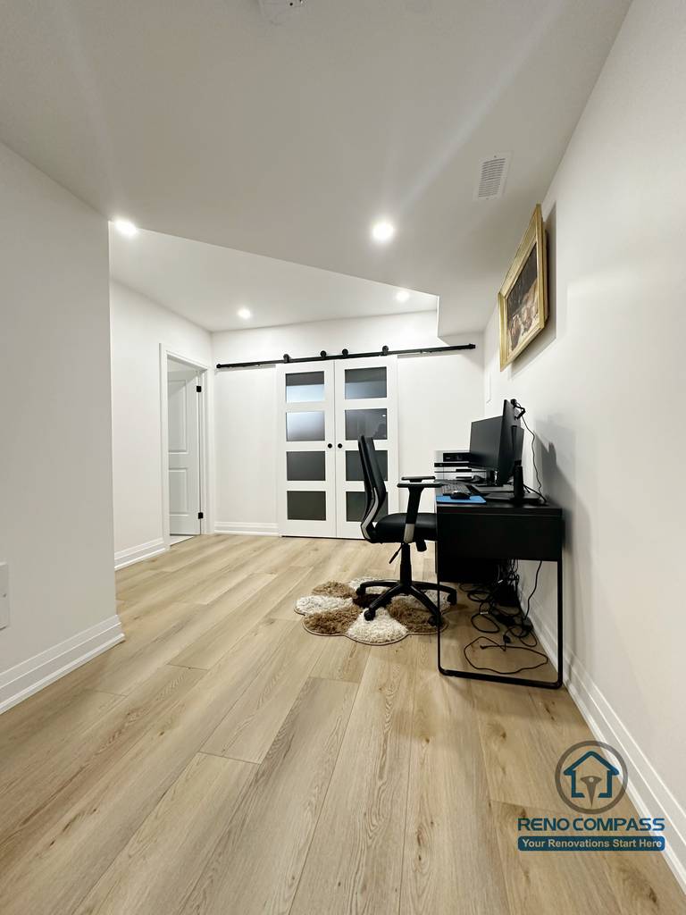 This Richmond Hill basement renovation features a flexible home office area designed for remote work or study. The space includes wide-plank flooring, recessed ceiling lights, and clean white walls that keep the area bright and uncluttered. Sliding barn-style doors add visual interest while allowing separation between work and living zones. The layout comfortably fits a desk setup and can easily adapt into a study room, hobby area, or multi-purpose basement workspace.
