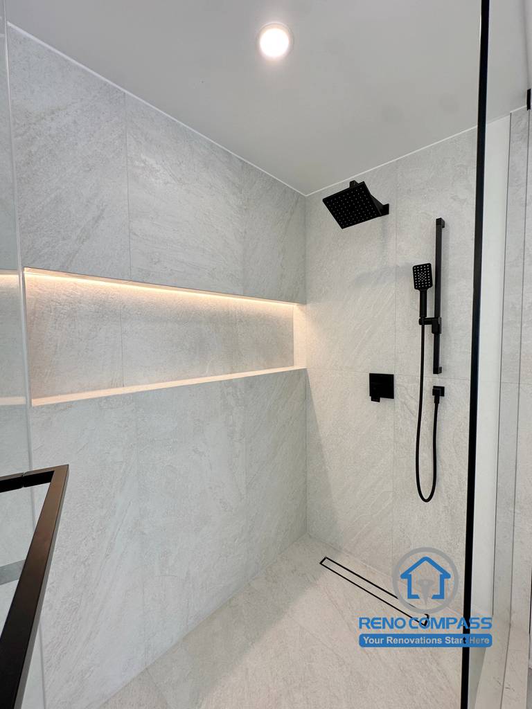 A luxury walk-in shower featuring frameless glass, illuminated recessed shower niches, matte black rainfall fixtures, linear floor drain, and large-format porcelain wall tiles. Completed during a whole home renovation in Markham, Ontario, this modern shower design delivers a spa-inspired experience with premium materials and expert craftsmanship by Reno Compass.