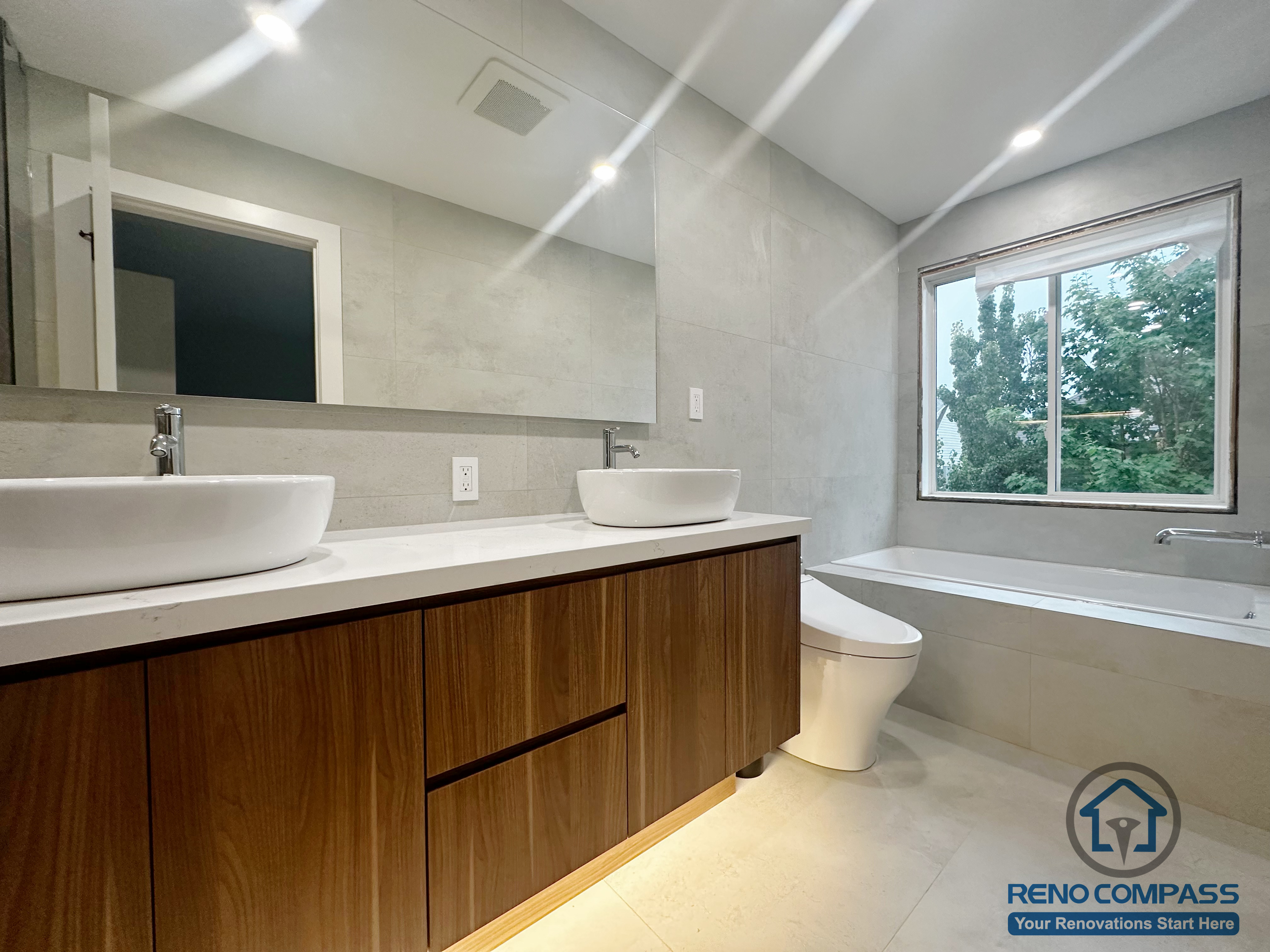 This Oakville bathroom renovation features a custom wood vanity with double vessel sinks, a full-width mirror, and soft recessed lighting. Neutral wall tiles and clean lines create a calm, modern atmosphere, while the window brings in natural light to enhance everyday comfort and functionality.