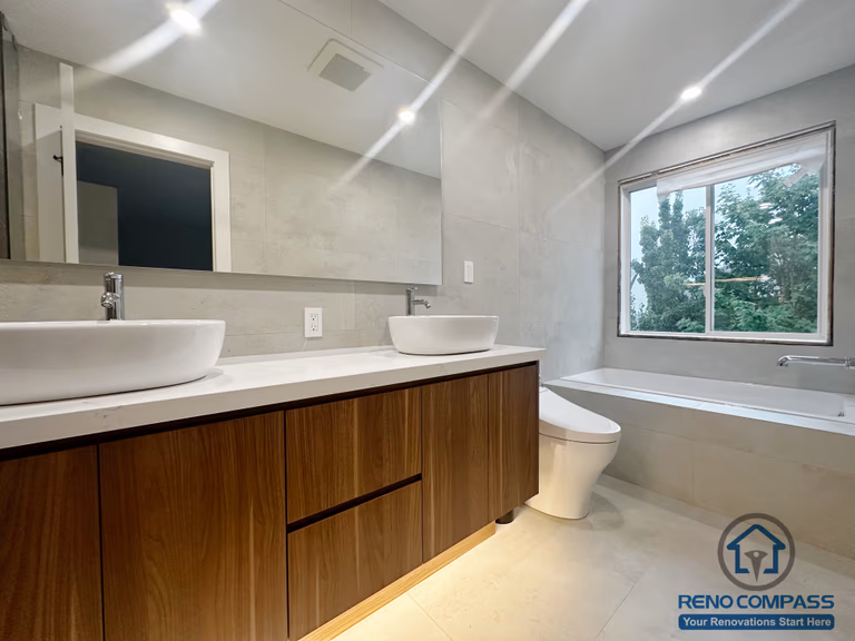 This Oakville bathroom renovation features a custom wood vanity with double vessel sinks, a full-width mirror, and soft recessed lighting. Neutral wall tiles and clean lines create a calm, modern atmosphere, while the window brings in natural light to enhance everyday comfort and functionality.