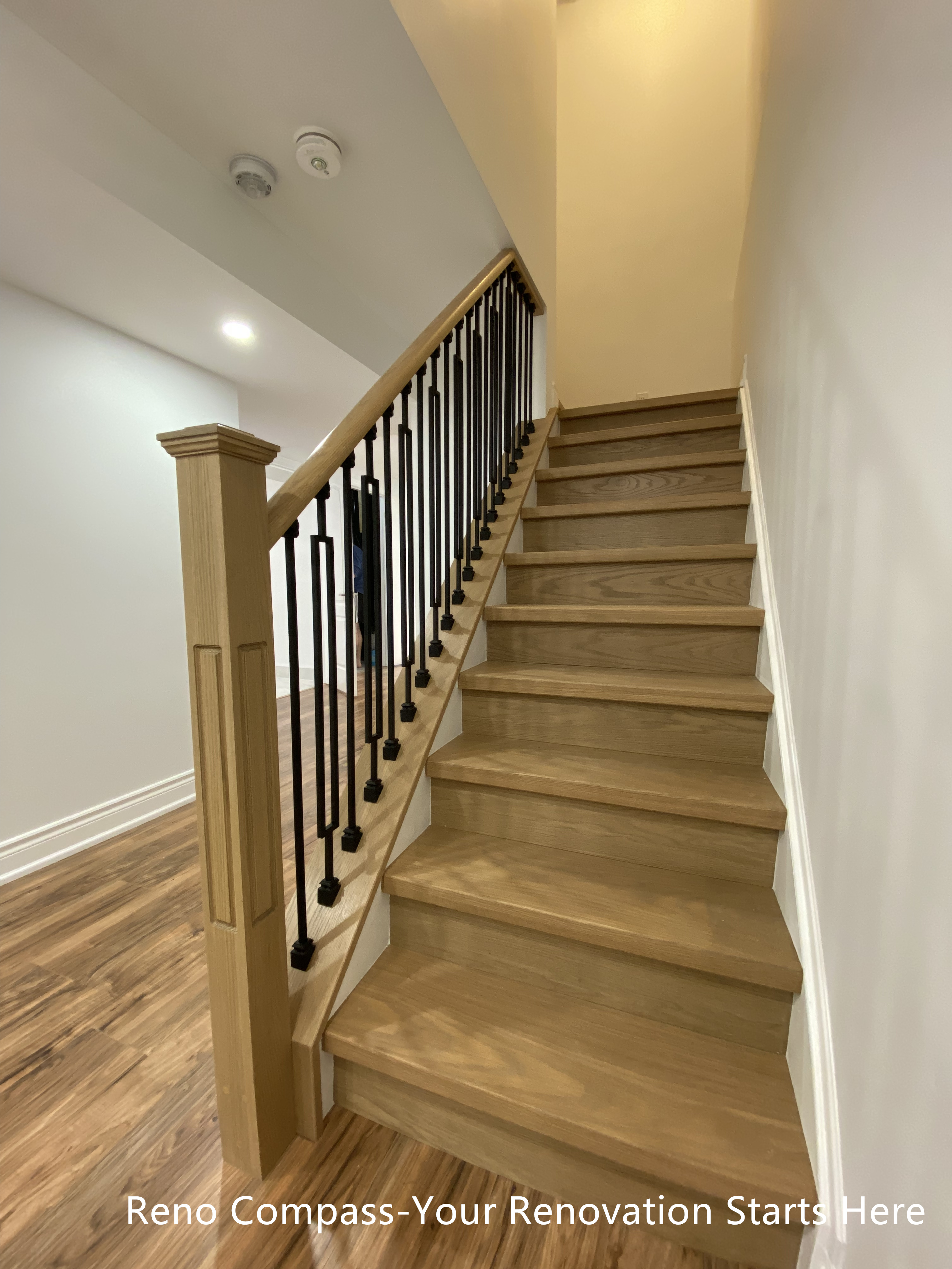This image showcases the newly renovated basement staircase in a Bradford basement renovation project, completed by Reno Compass Inc. The staircase features solid wood treads with a warm natural finish, paired with black iron balusters and a classic square newel post for added durability and visual balance. The open railing design improves sightlines and allows light to flow through the basement, making the space feel brighter and more connected. Durable flooring and recessed ceiling lighting complete the transition between levels, ensuring both safety and long-term functionality. This custom staircase renovation reflects a thoughtful approach to basement layout, circulation, and everyday usability for Bradford homeowners.