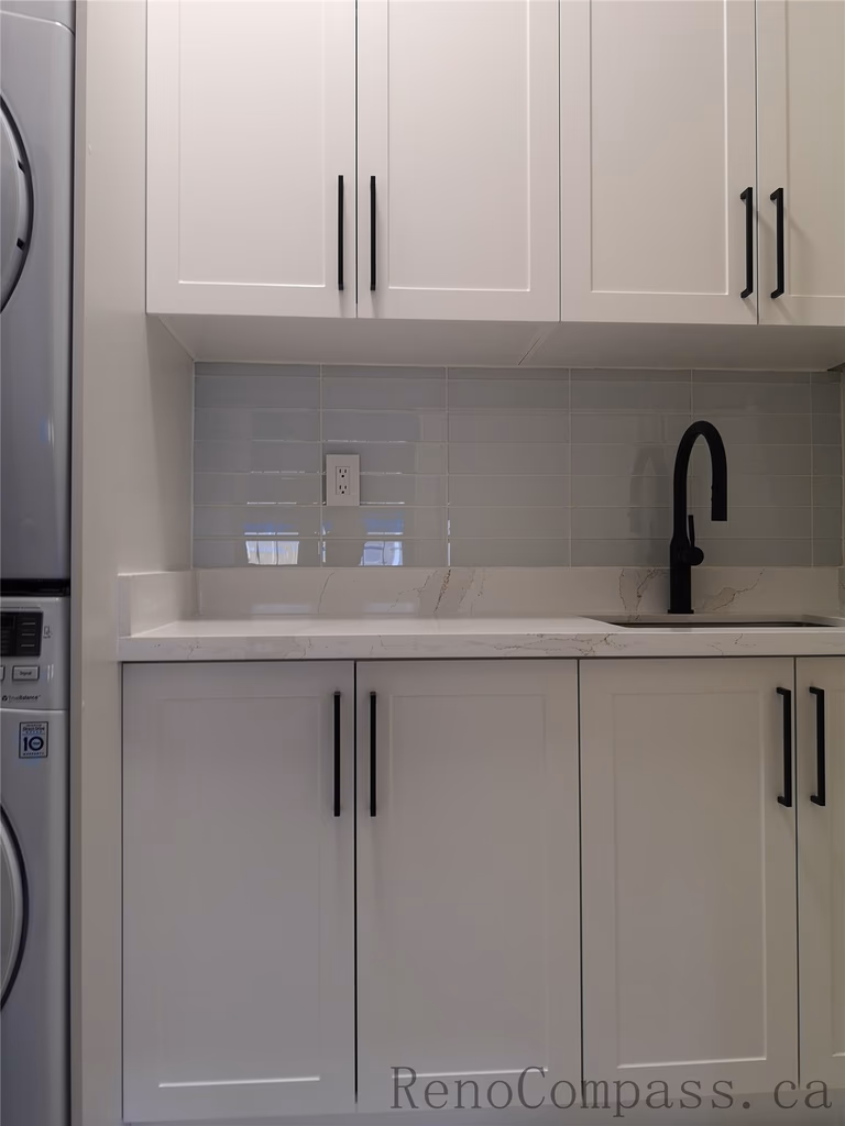 This Aurora home renovation features a clean and modern laundry room design with custom white shaker cabinets, quartz countertop, and a built-in utility sink with matte black faucet. The stacked washer and dryer layout maximizes space efficiency while maintaining a bright and organized look. Completed by a professional Toronto renovation company focused on practical storage solutions, modern finishes, and high-quality craftsmanship.