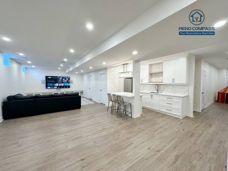 This image showcases the open-concept basement layout in a Bradford basement renovation project, featuring a seamless connection between the living area and the custom kitchenette. White cabinetry, a functional peninsula with seating, and recessed ceiling lights create a bright and welcoming space. The continuous flooring enhances visual flow, making the basement suitable for family gatherings, entertainment, and everyday use. Designed and built by a Toronto renovation company with a focus on comfort, durability, and practical space planning.