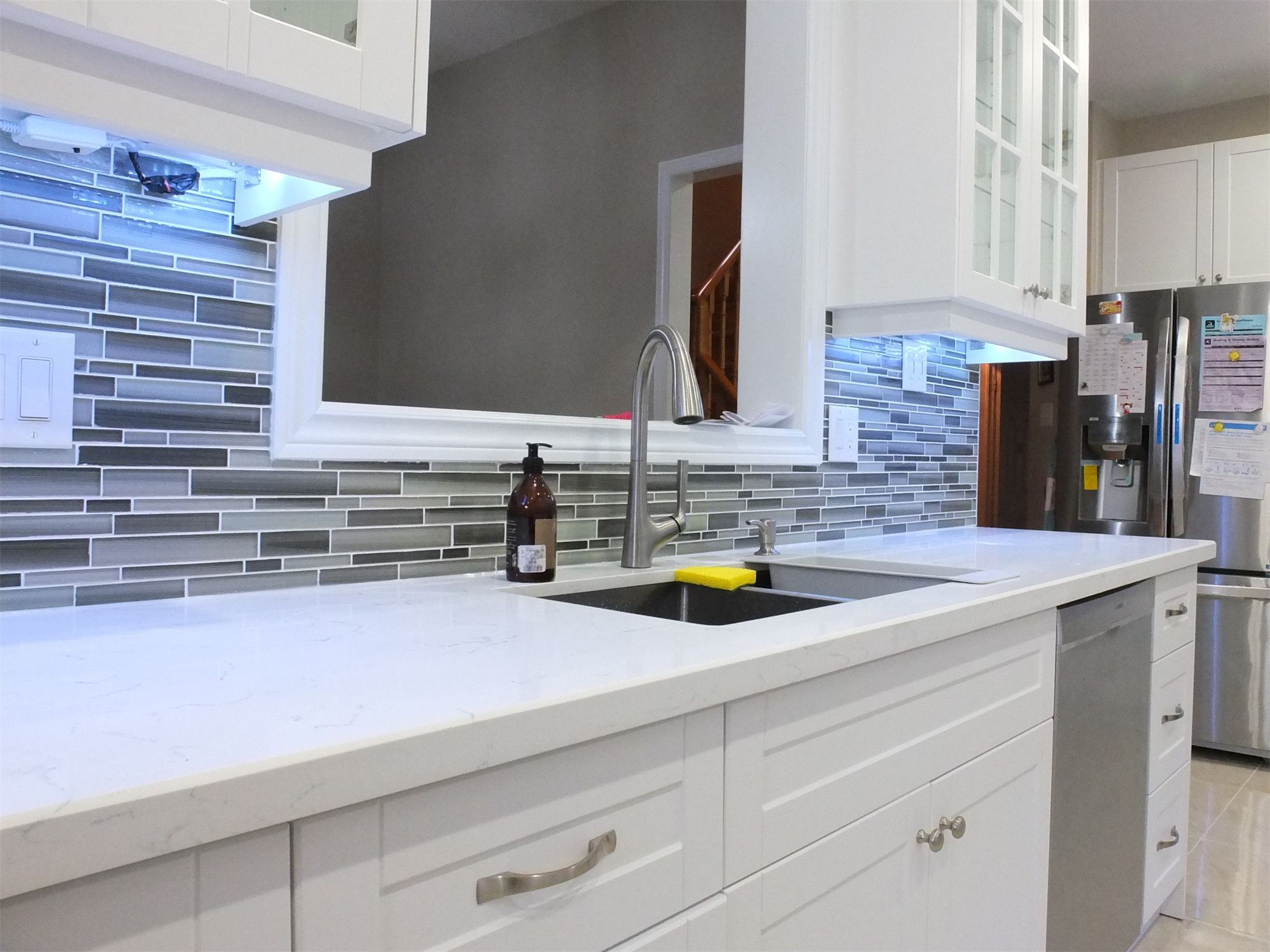 This Vaughan kitchen renovation highlights elegant white shaker cabinets with glass display doors, quartz countertops, and a modern glass mosaic backsplash enhanced by LED under-cabinet lighting. The clean layout and bright finishes create a stylish and functional cooking space. Completed by a trusted Toronto renovation company, the design blends modern aesthetics with everyday practicality.