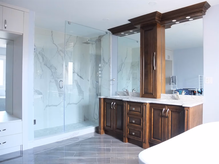 This Richmond Hill bathroom renovation features a modern glass walk-in shower with marble-style wall tiles, a custom wood vanity with quartz countertop, and upgraded lighting. The design combines luxury finishes with practical storage to create a bright and elegant bathroom space.