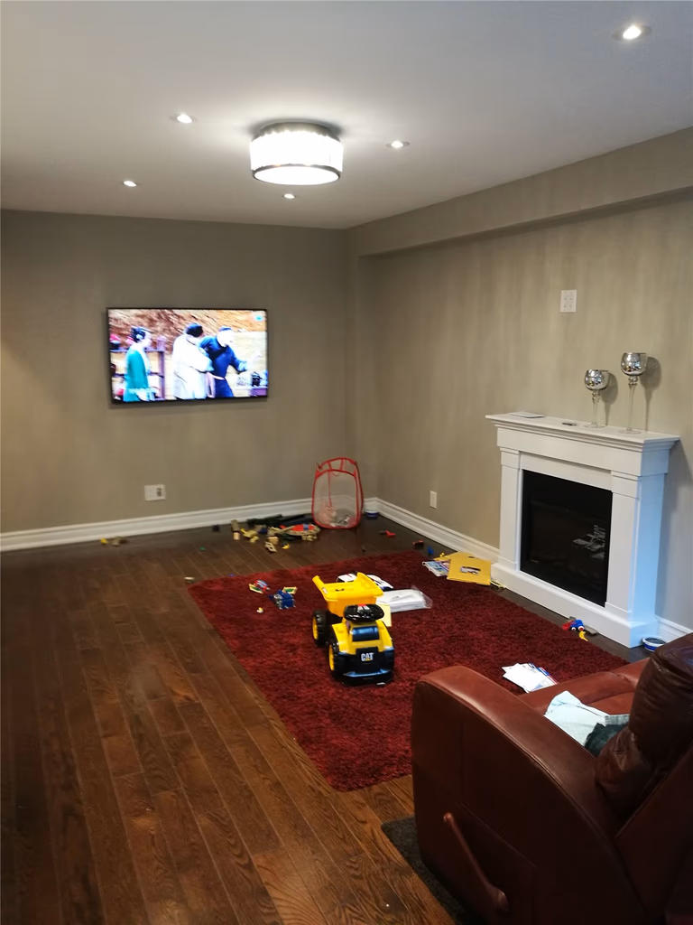 This Newmarket interior renovation features a refreshed family room with dark hardwood flooring, recessed ceiling lights, and a modern flush-mount light fixture. A clean fireplace surround and wall-mounted TV create a comfortable and functional living space, showcasing the quality craftsmanship of a professional Toronto renovation company.